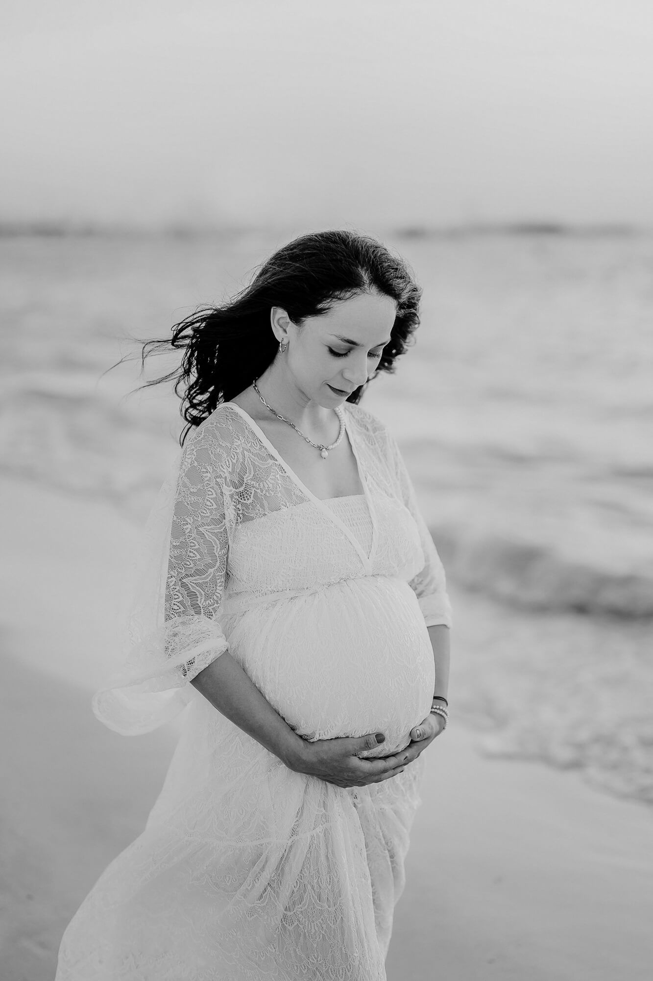 A pregnant person in a white lace dress stands thoughtfully by the ocean in a black and white photograph.