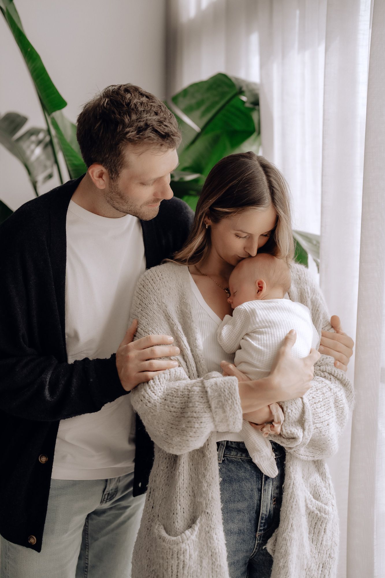 A family in cozy knit sweaters shares an intimate moment with their baby near a large tropical plant by a window.