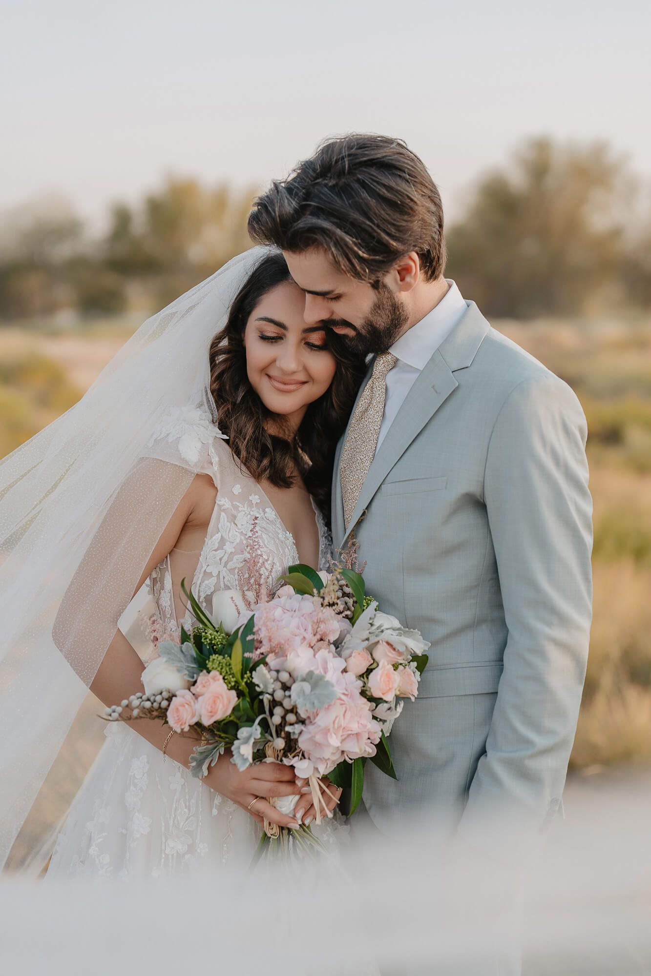 A romantic wedding portrait showcases a tender moment between newlyweds with a soft pink bouquet in a rustic outdoor setting.