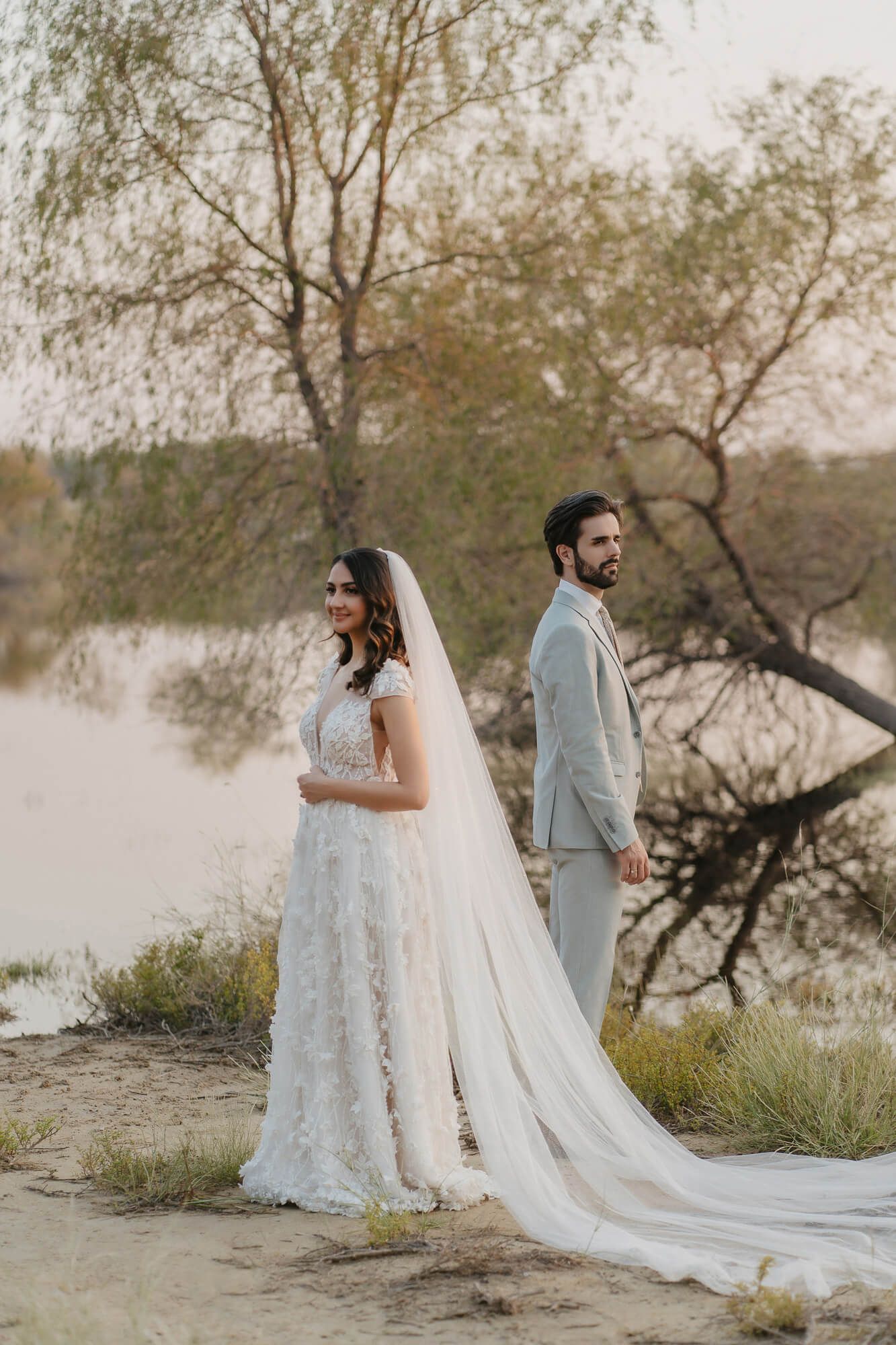 A couple stands back to back by a lake at sunset, with bride wearing a flowing lace gown and long veil.