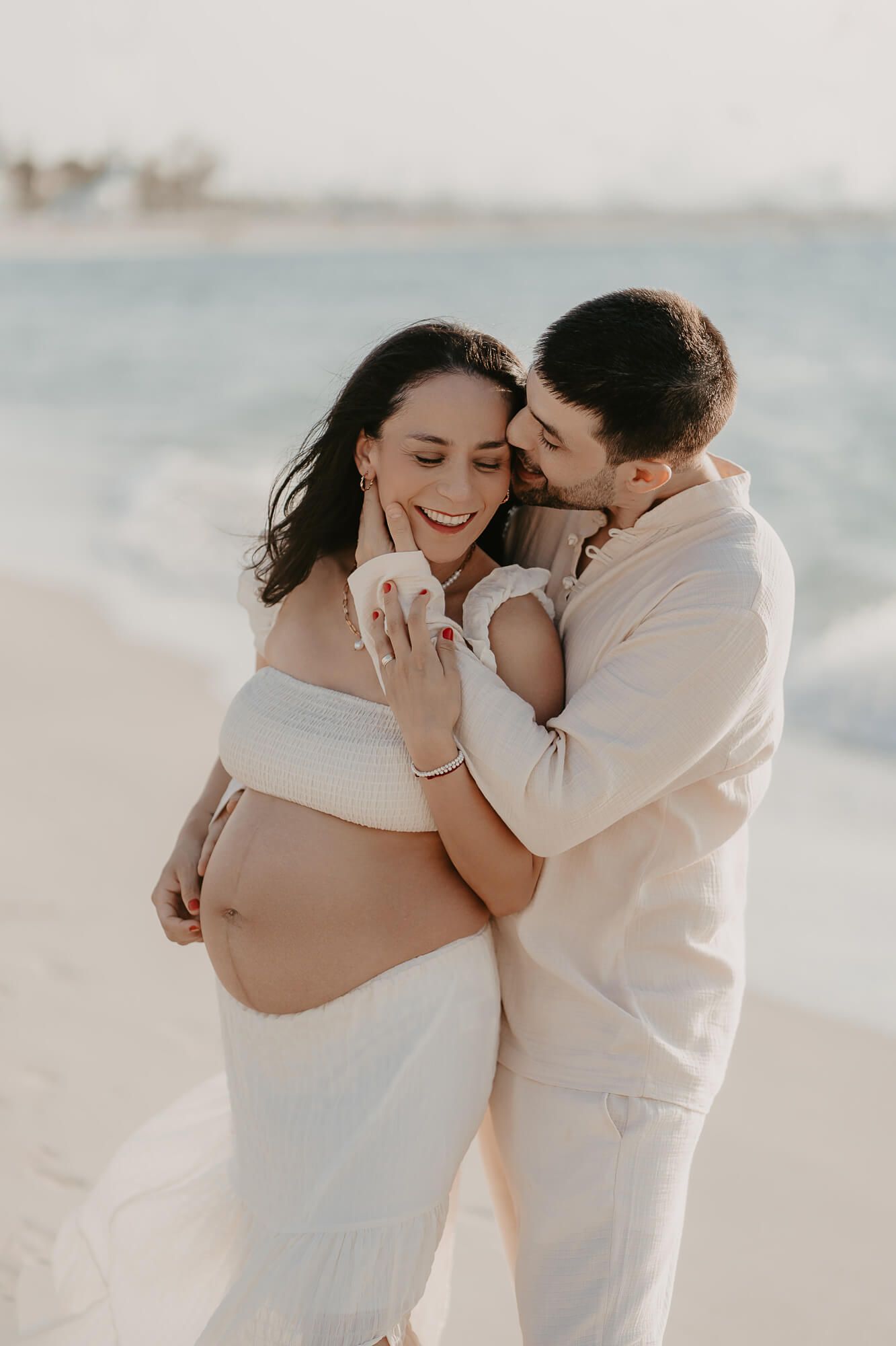 Romantic couple in white attire embraces on beach during maternity photoshoot with ocean in background.