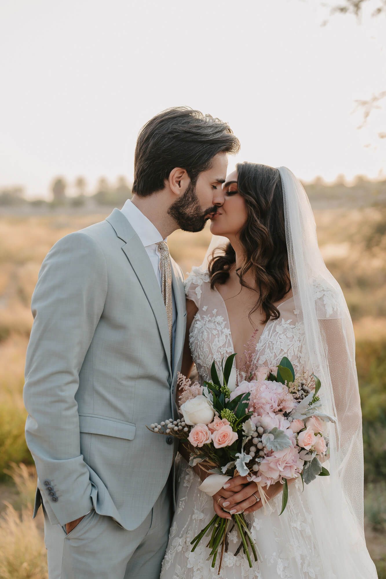 A romantic wedding couple embrace in a golden field at sunset while the bride holds a beautiful pink bouquet.