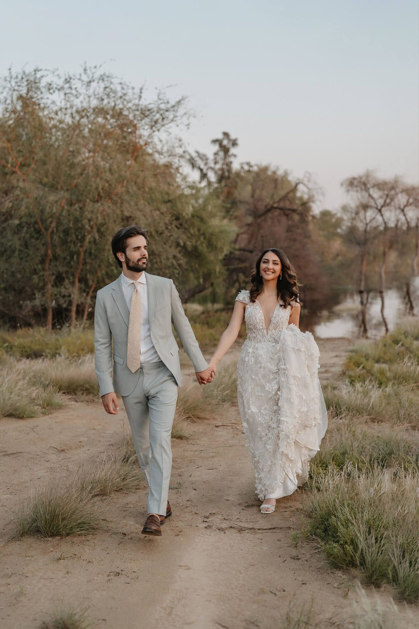A couple walks hand in hand on a dirt path wearing wedding attire against a natural desert landscape at sunset.