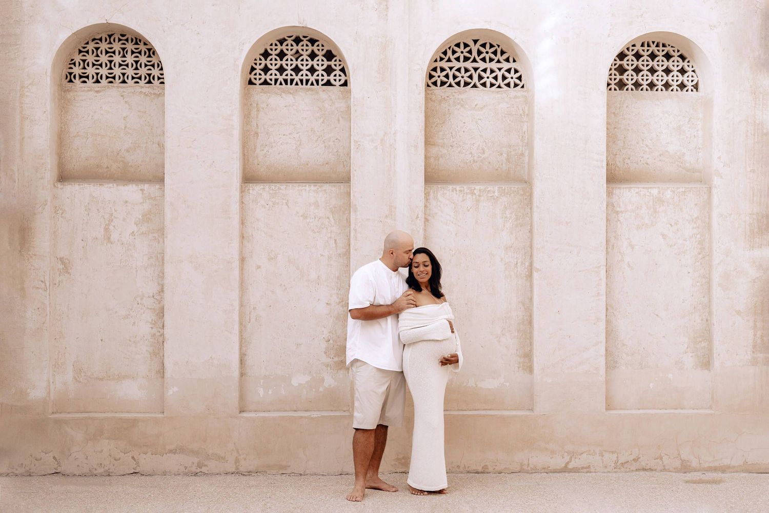 A couple poses for a maternity photo against a wall with decorative arched alcoves.