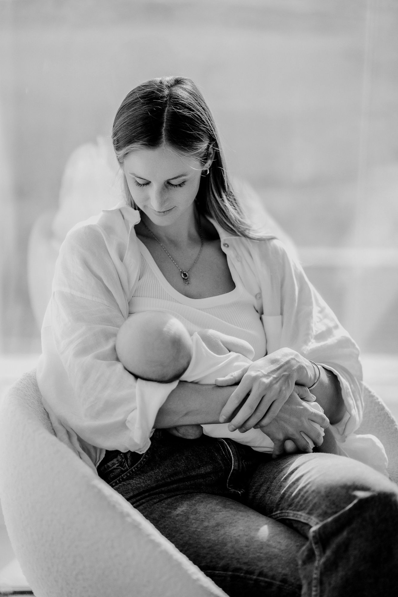 A tender black and white portrait of a nurturing mother in jeans and white cardigan breastfeeding her newborn baby.