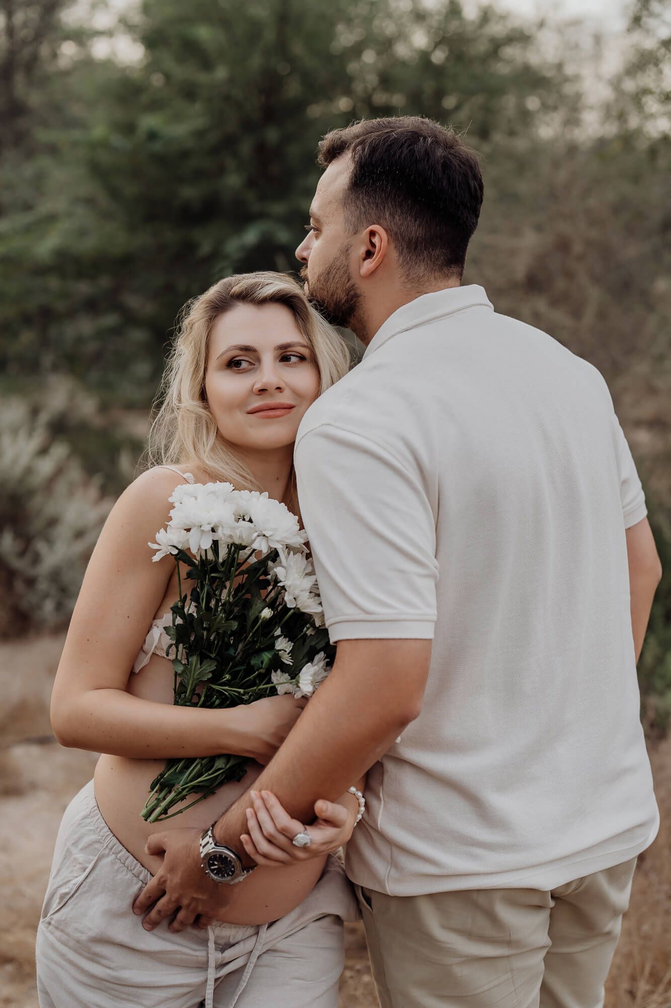 A romantic couple embraces while holding white flowers against a natural outdoor backdrop.