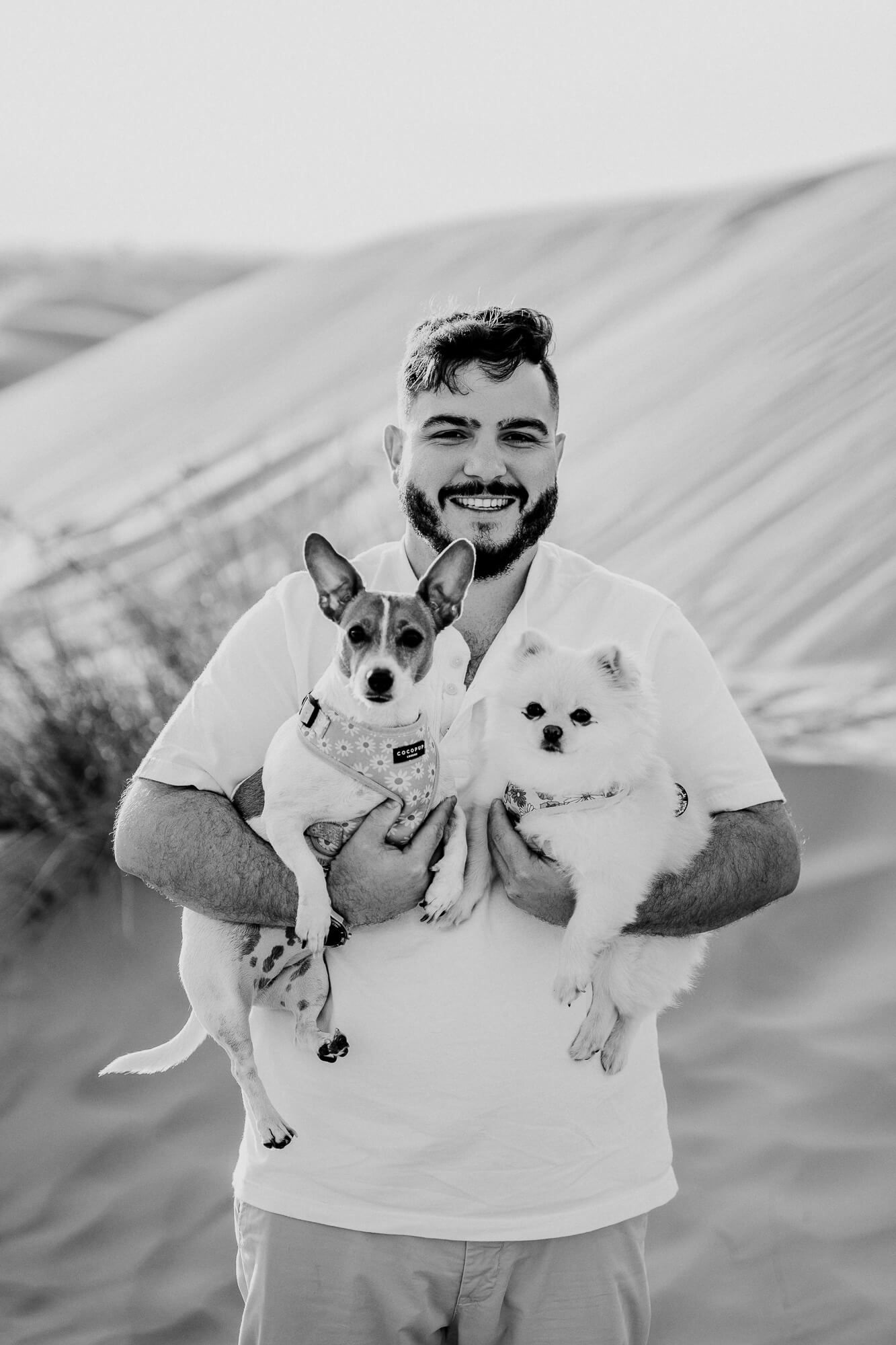 Person holding two small dogs, a Jack Russell mix and white Pomeranian, against sandy desert background.
