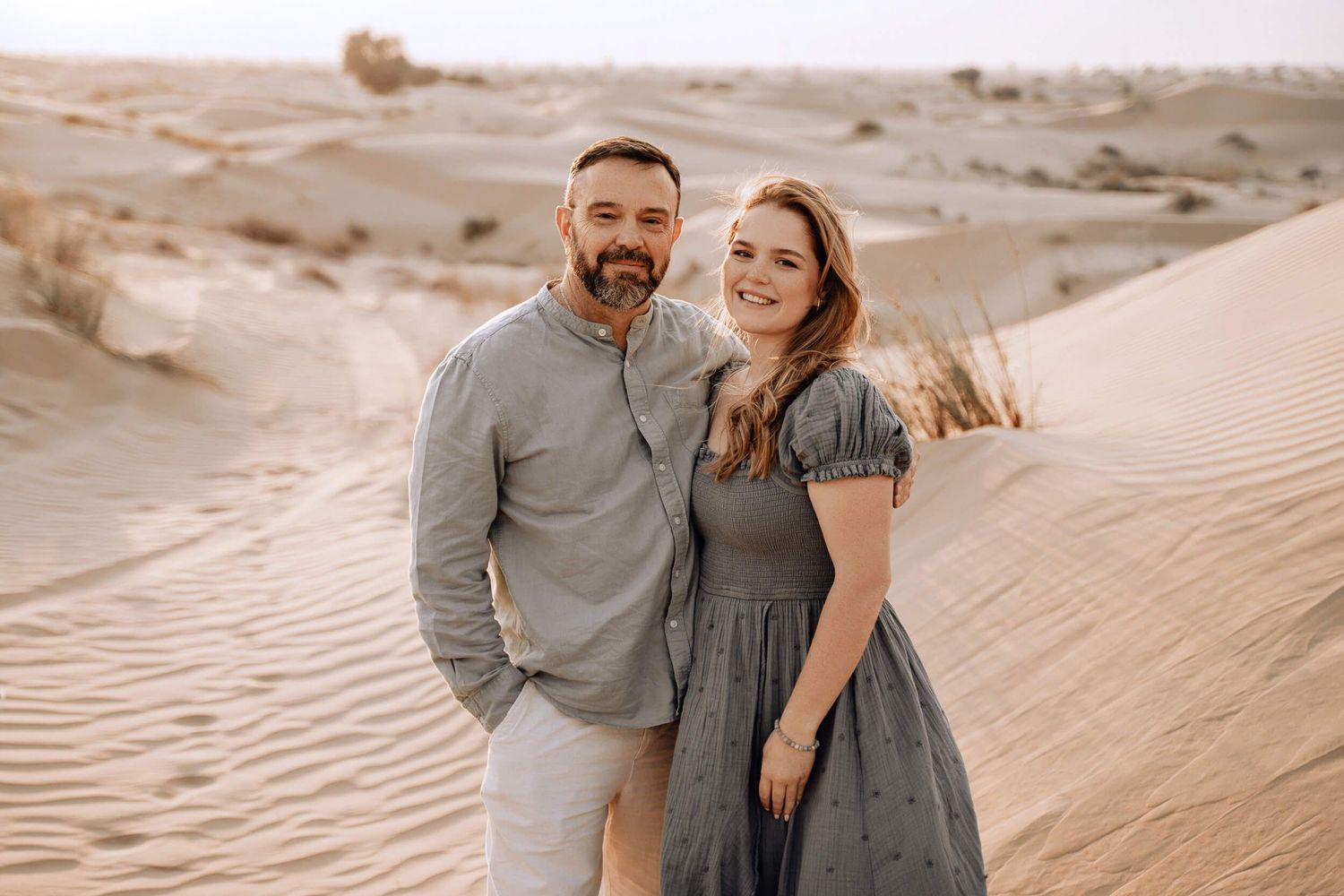 A romantic desert photoshoot featuring a couple standing on sand dunes in complementary outfits.