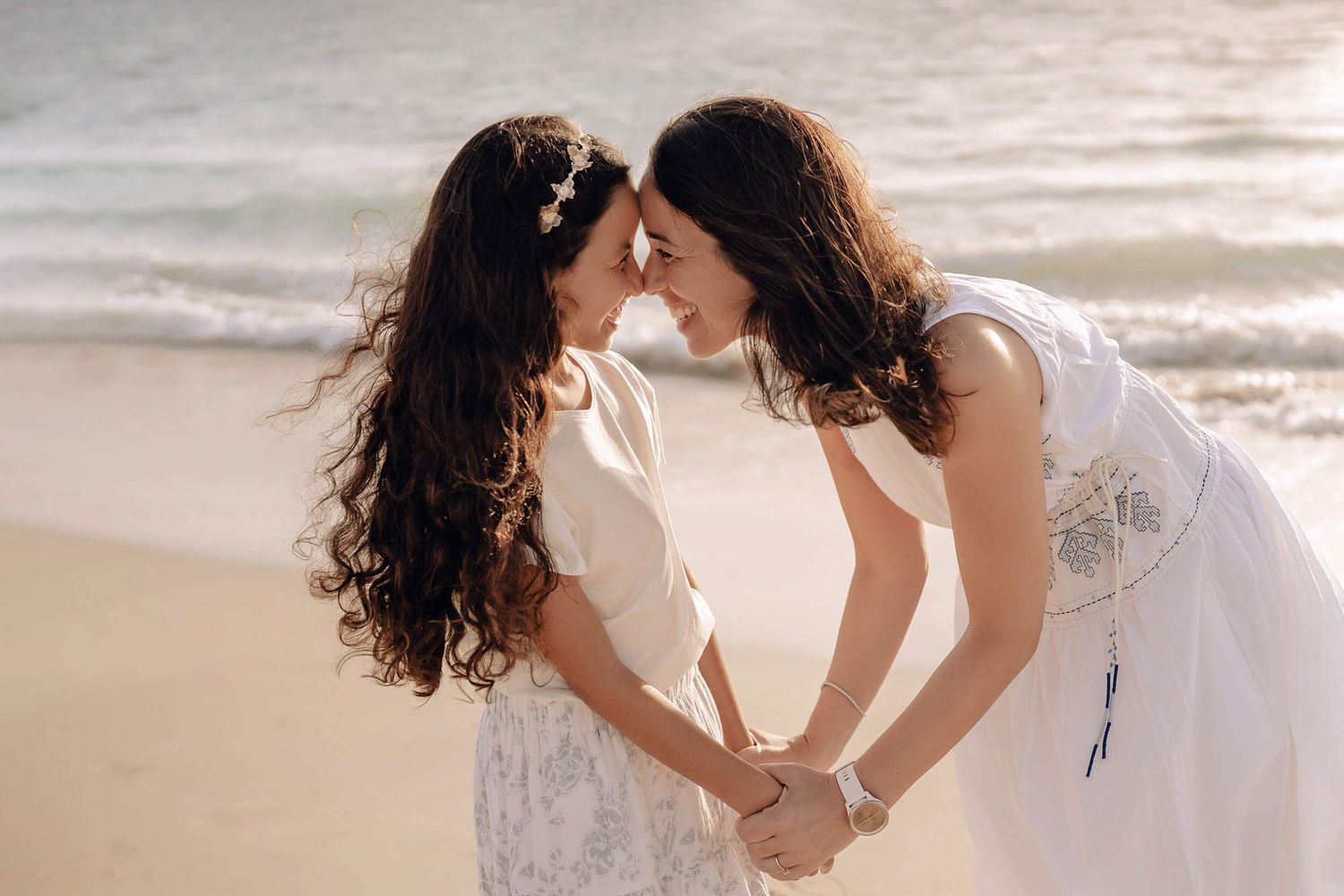 Two people in white dresses share an intimate moment while holding hands on a sunny beach.