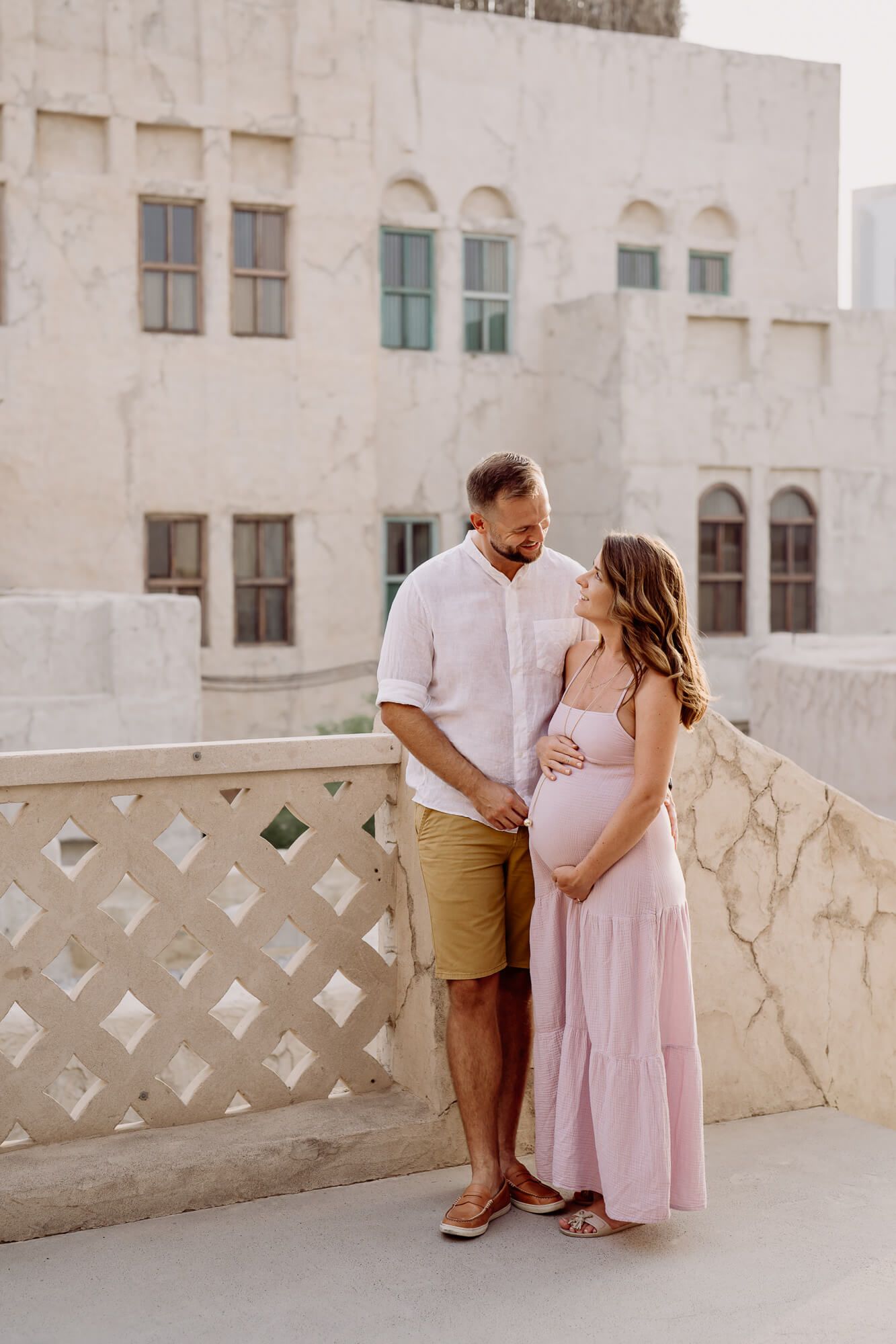 Expectant couple in light summer outfits standing together on a balcony with white architectural details in the background.