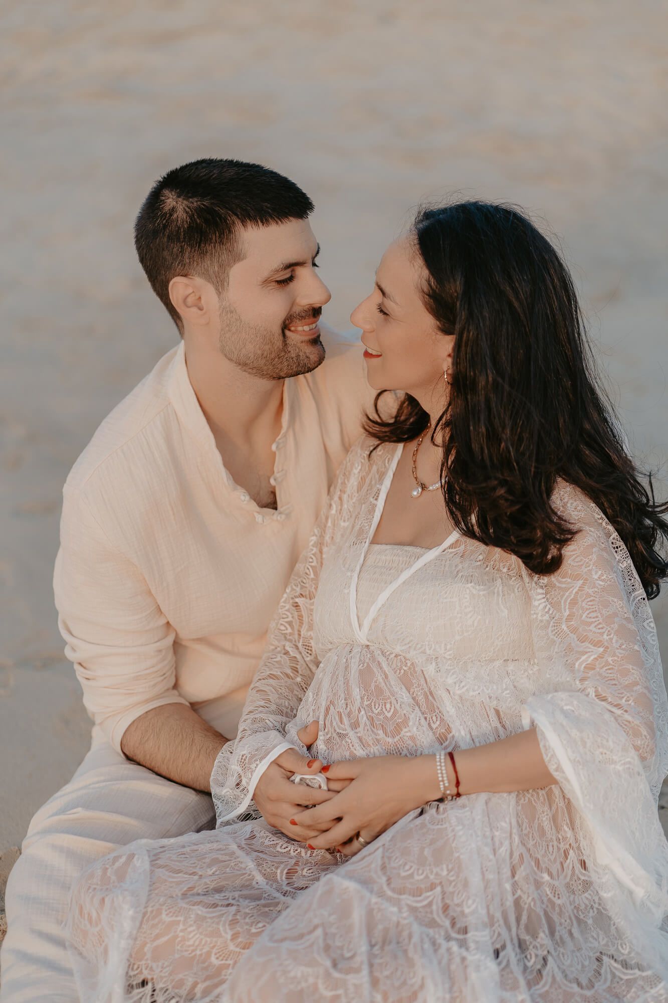 A romantic maternity session features a couple in white outfits sitting together against a muted backdrop at sunset.