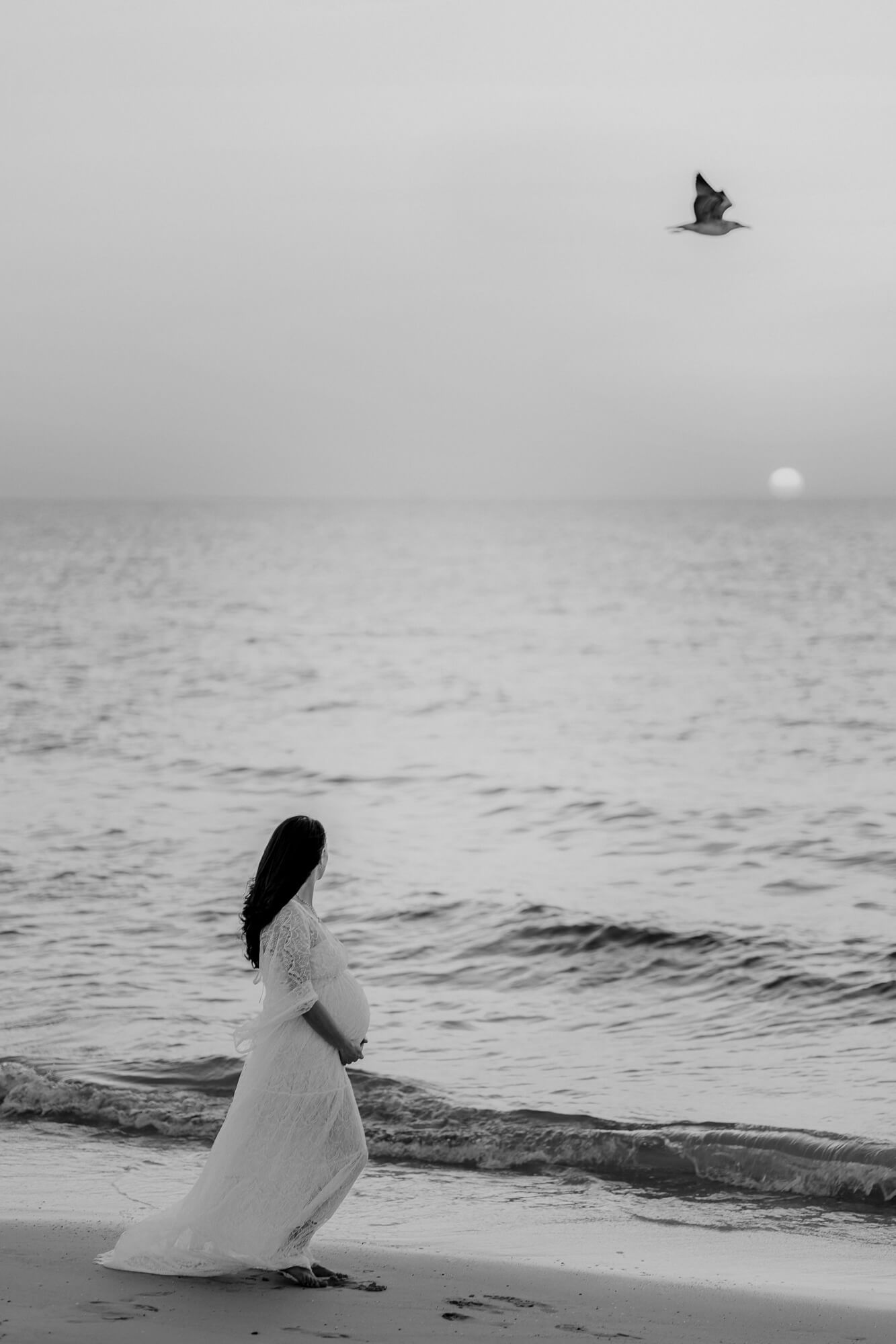 A minimalist black and white photo series captures an expectant mother in white on a beach at sunset with birds soaring above.