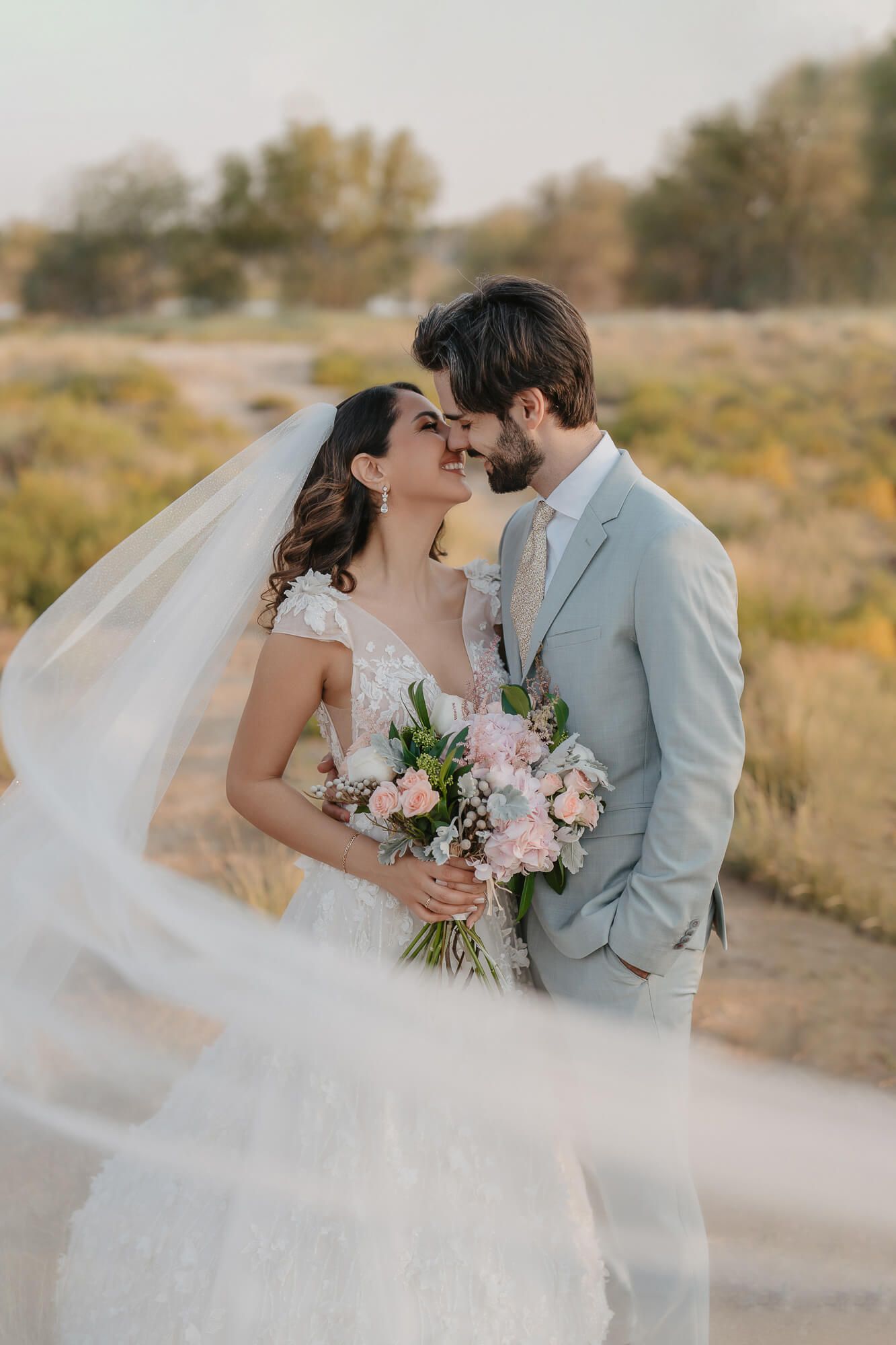 A couple shares an intimate moment in a golden field holding a pink bouquet while wearing wedding attire.
