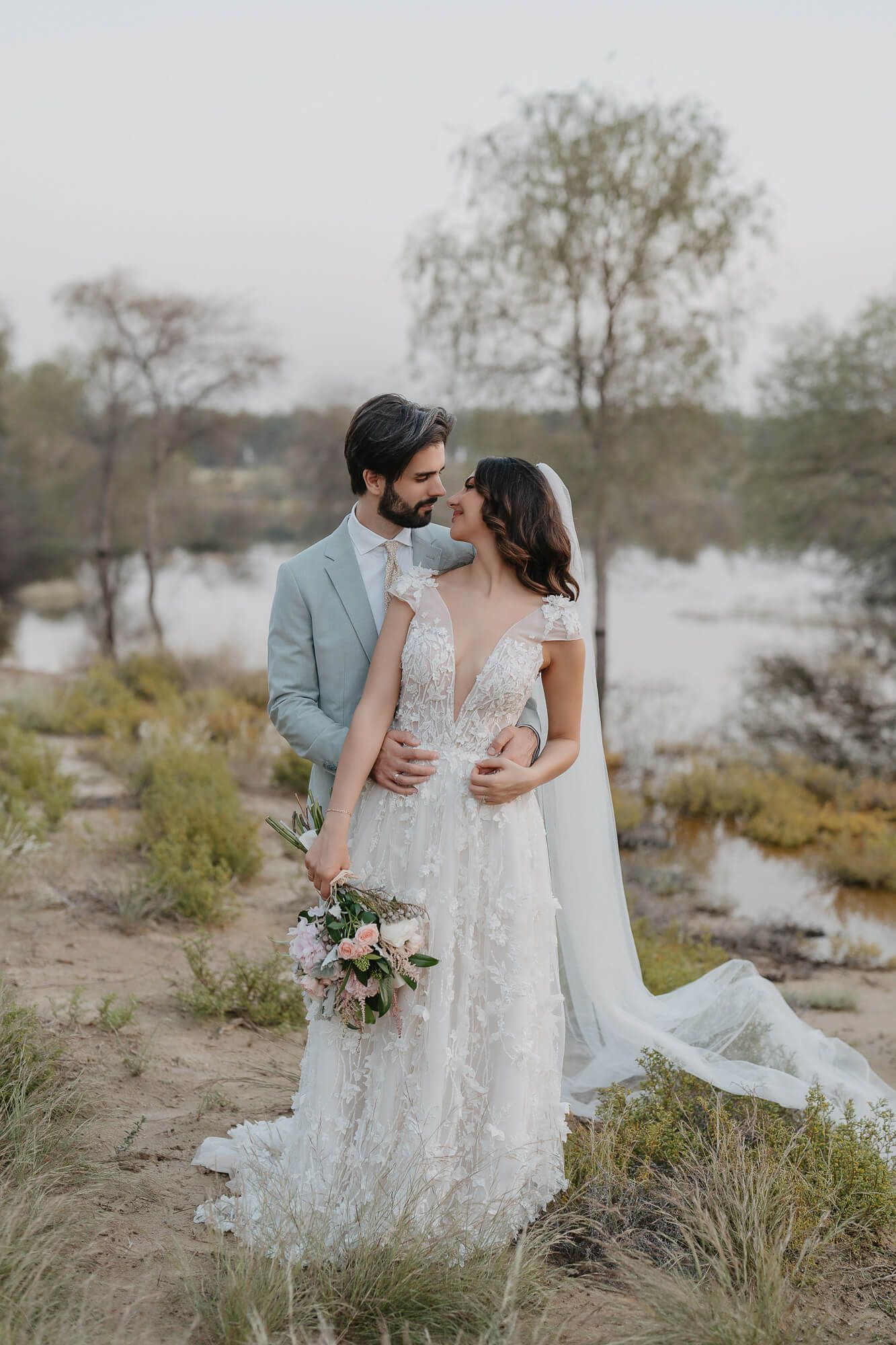 A romantic wedding couple embraces in a rustic outdoor setting with flowing white dress and natural scenery.