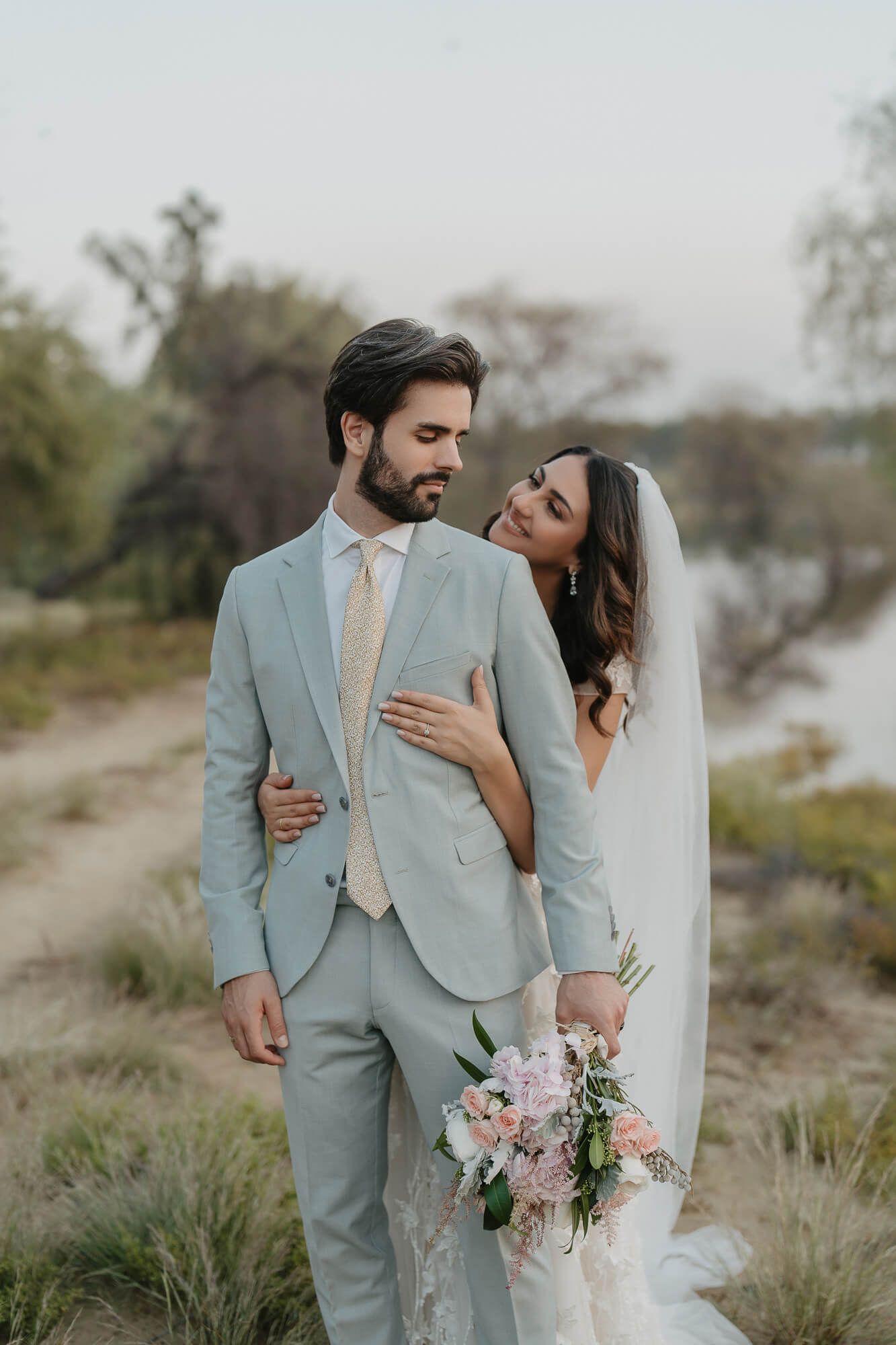 Intimate outdoor desert wedding photos with couple in soft sage green attire and delicate pink bouquet amid natural scenery.