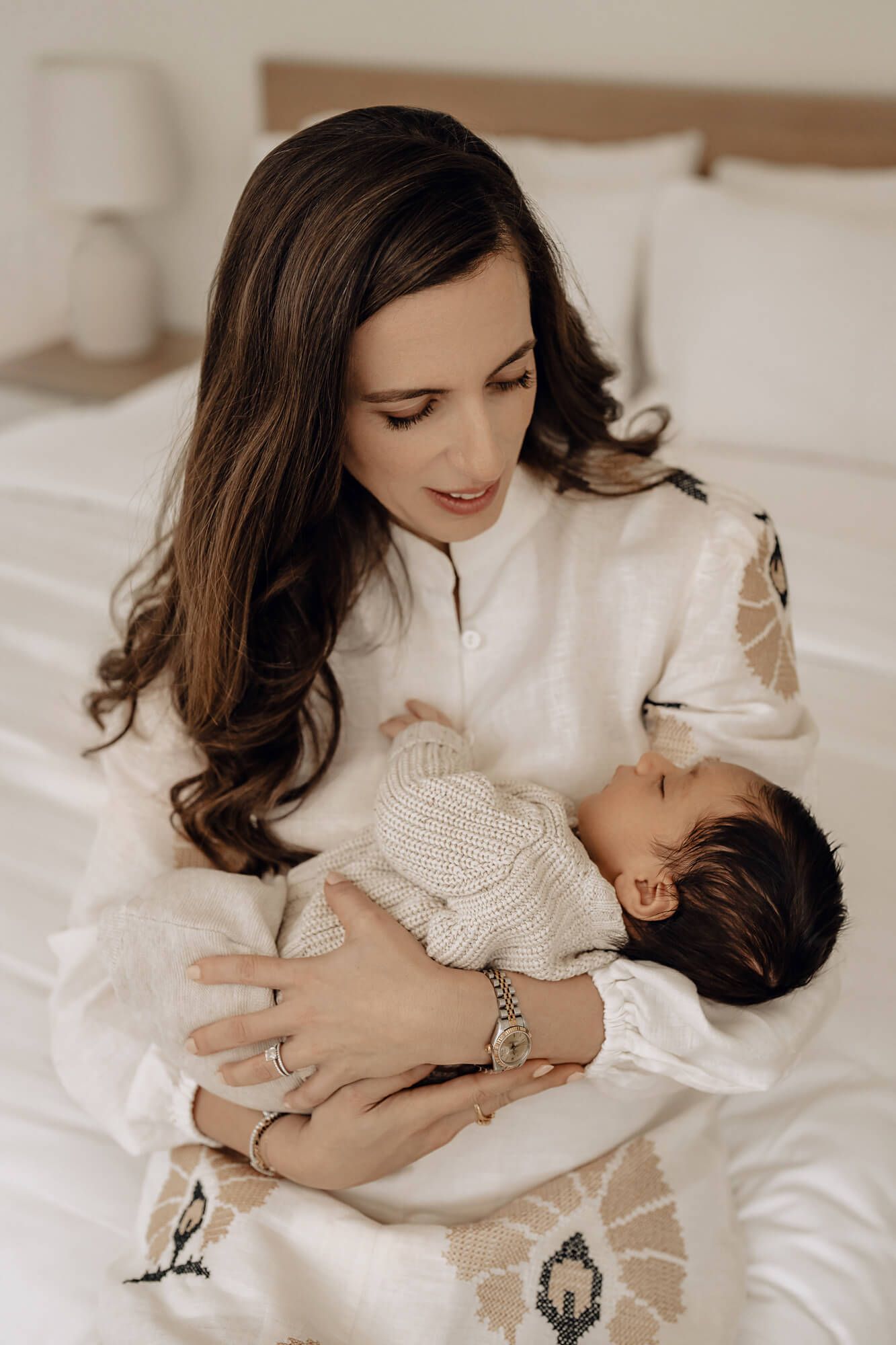 A woman in a white blouse cradles a sleeping baby wearing a knit sweater while sitting on white bedding.