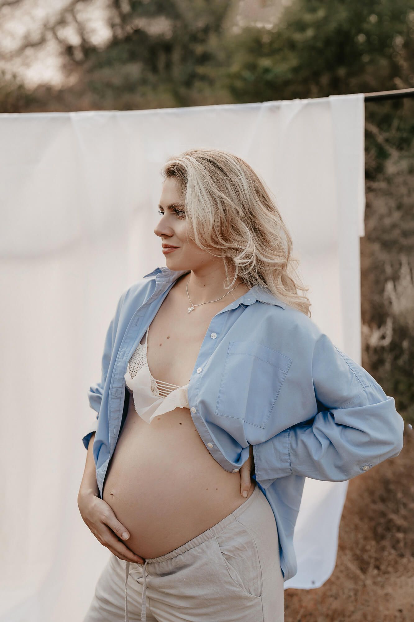 A maternity photoshoot against a white backdrop features a casual blue button-up shirt and white bralette outfit.