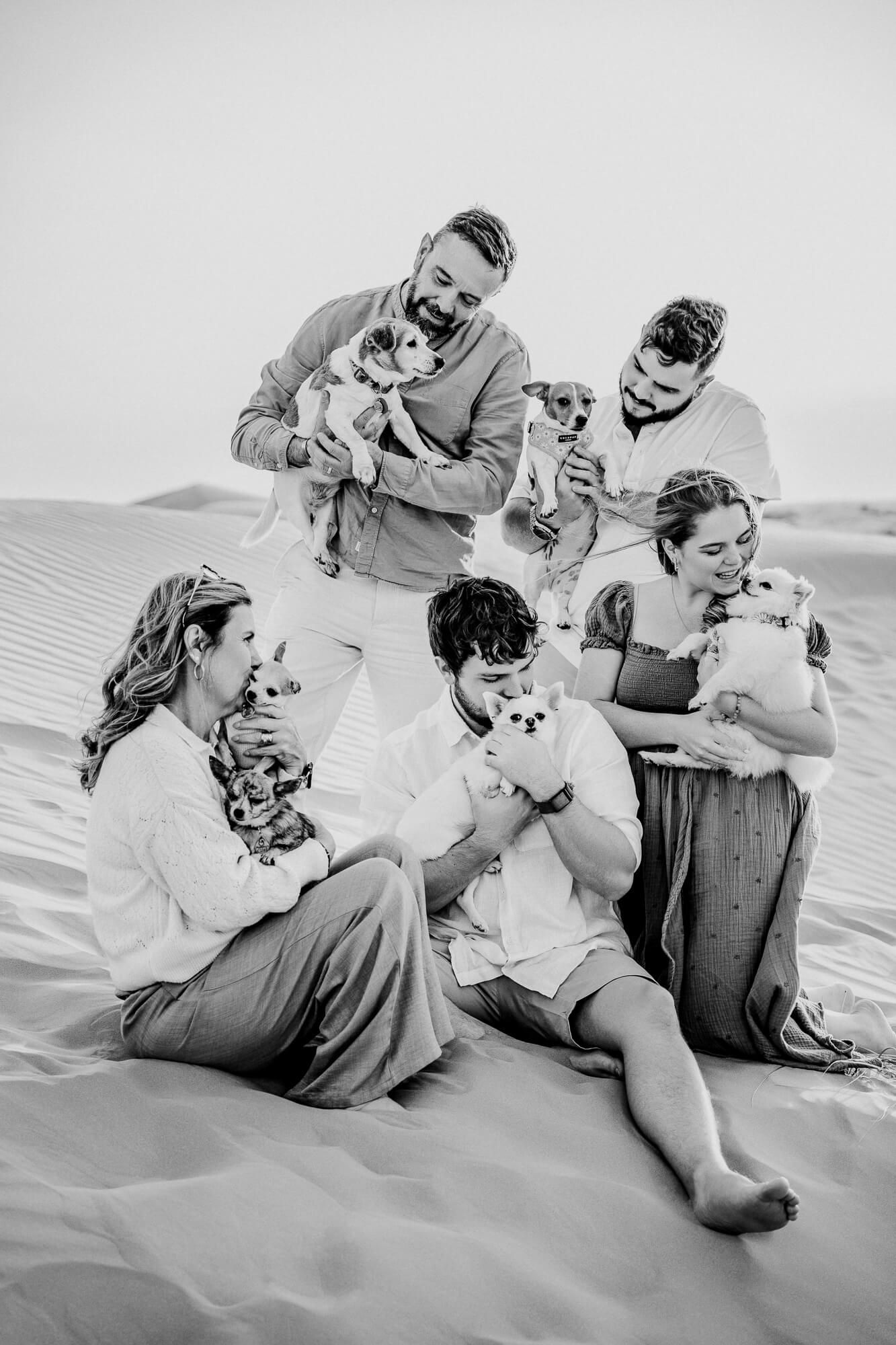 Black and white photo of five people cuddling with their small dogs on desert sand dunes.