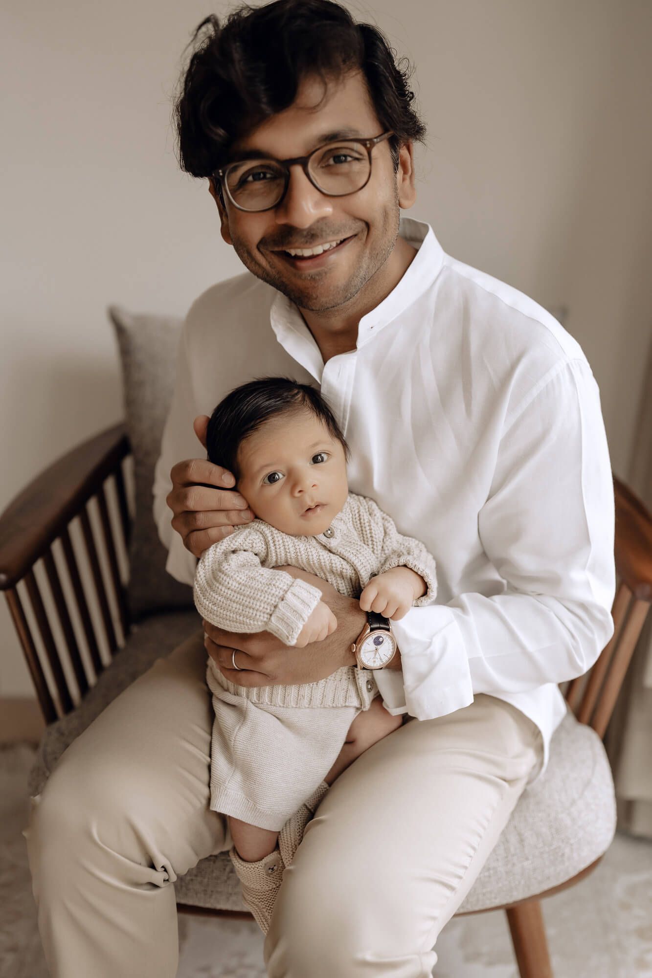 Parent and baby in matching white outfits sitting on wooden chair.