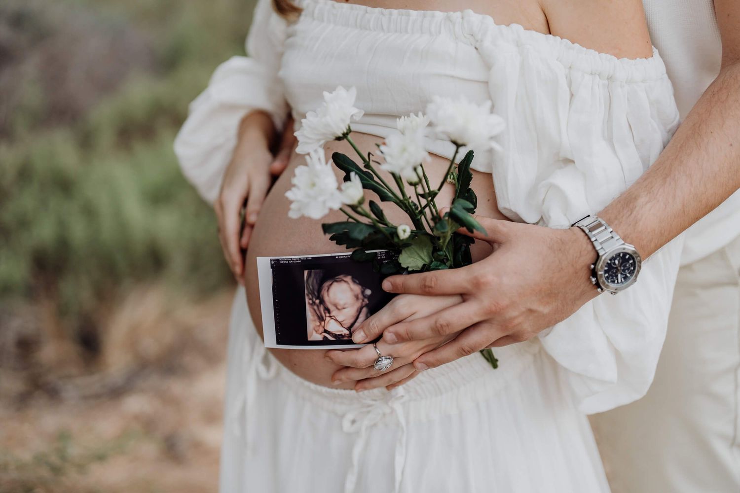 Close up of hands holding ultrasound photo, flowers and baby clothes against white dress.