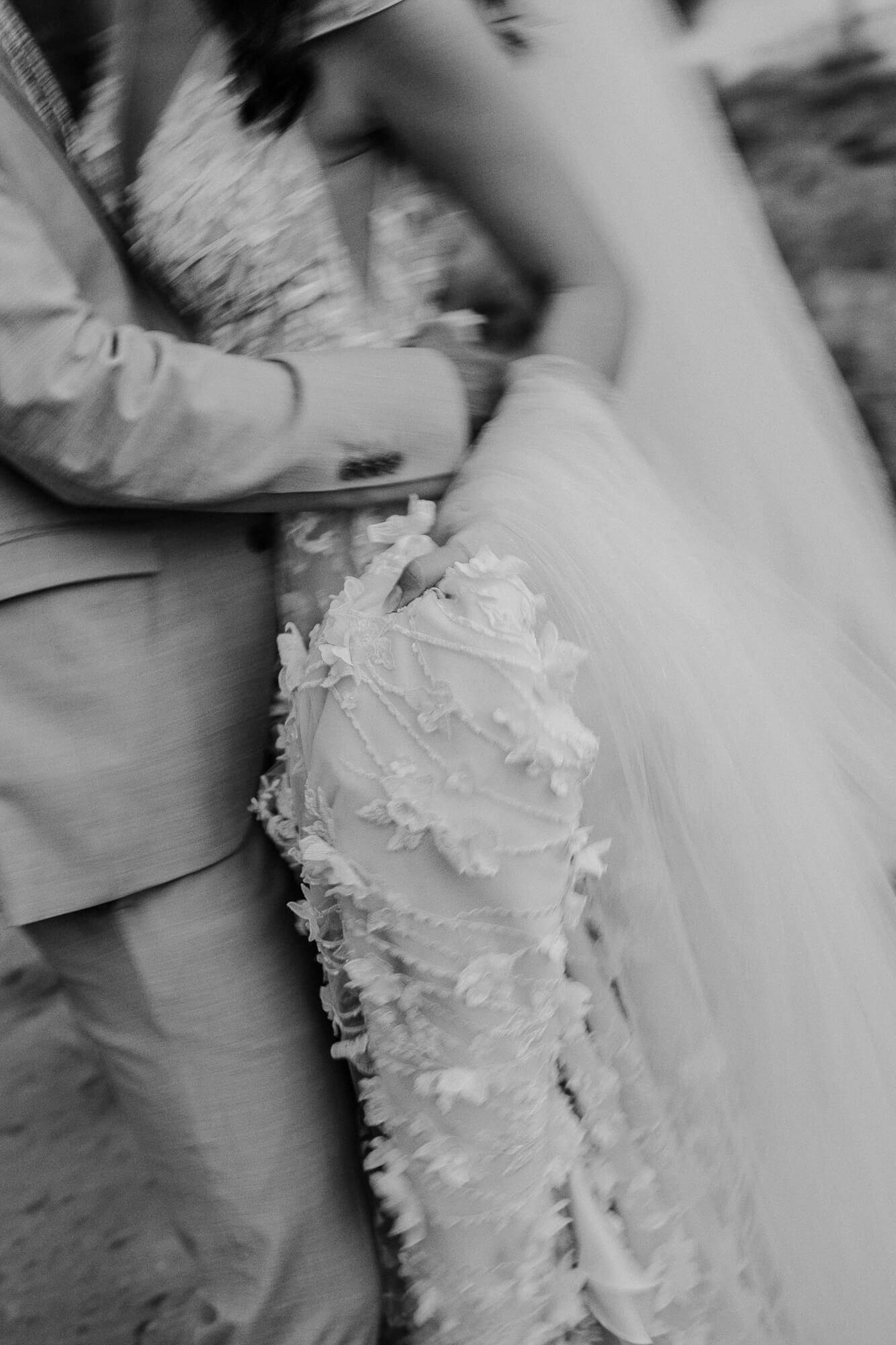 Black and white close-up photograph of wedding dress details with delicate lace and tulle fabric against a suit.