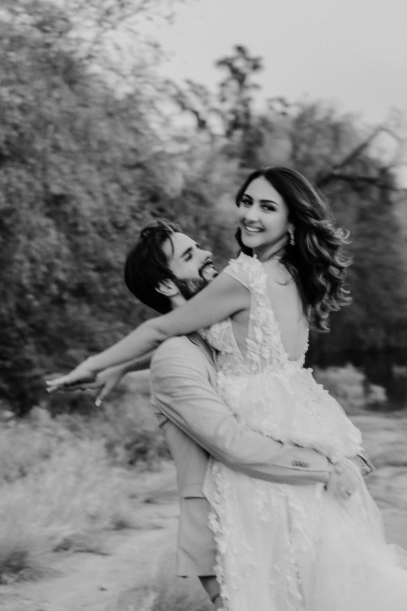 A romantic black and white portrait of a couple embracing joyfully outdoors, with the bride wearing a lace gown.