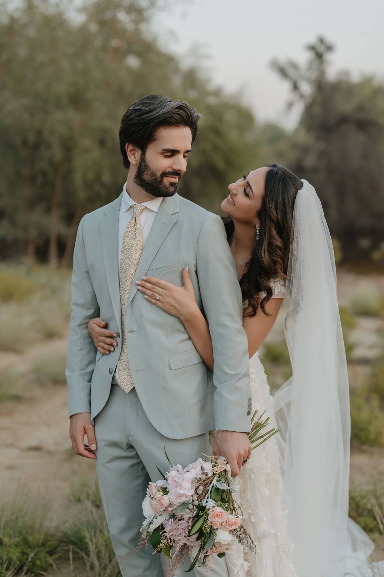 Newlyweds share an intimate moment outdoors as bride holds pink bouquet while wearing a lace gown and veil.
