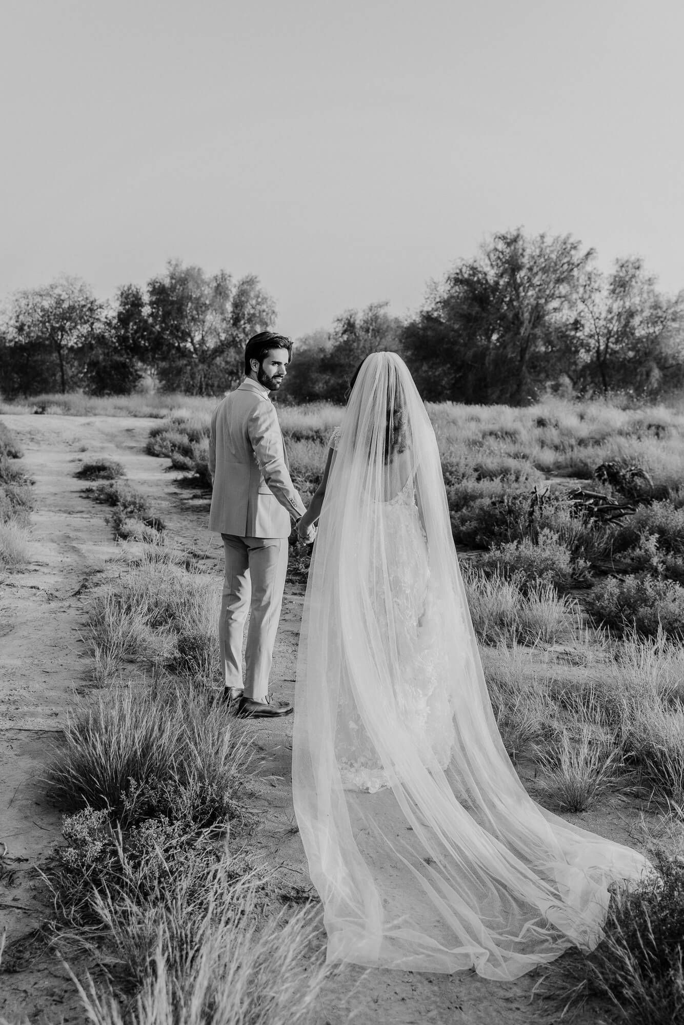 Black and white wedding photo of a couple standing on a desert path with a flowing long veil in the foreground.