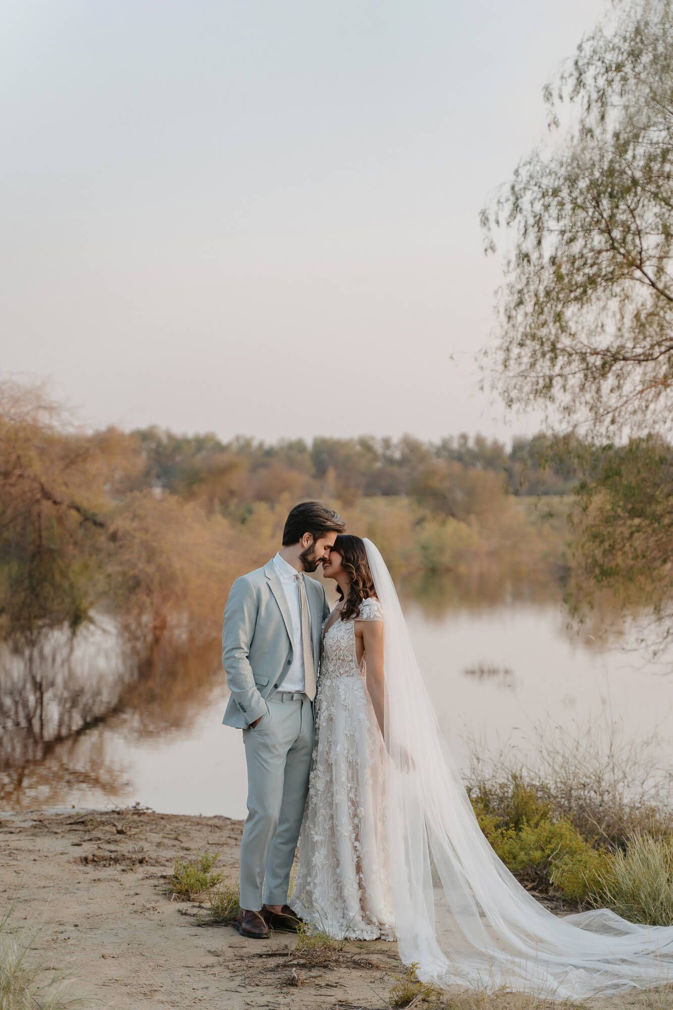 A tender moment between newlyweds in formal attire standing by a serene lake during golden hour.