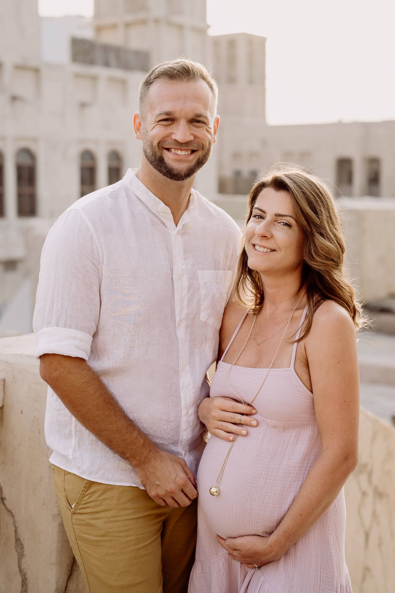 A couple in light coordinating outfits embraces during a maternity photoshoot with a bright architectural backdrop.