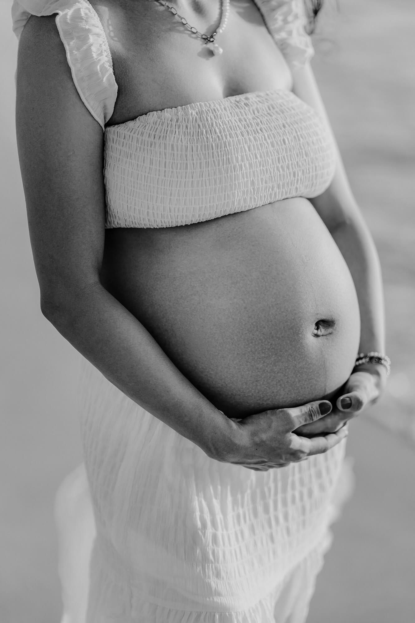 Black and white maternity photo showing a pregnant belly with a white bandeau top and flowy white skirt.