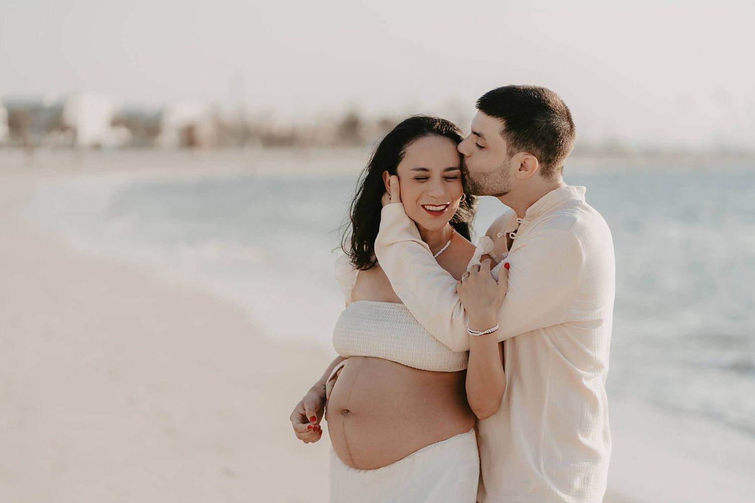 A couple shares a tender moment on a sandy beach during maternity photo session at sunset.