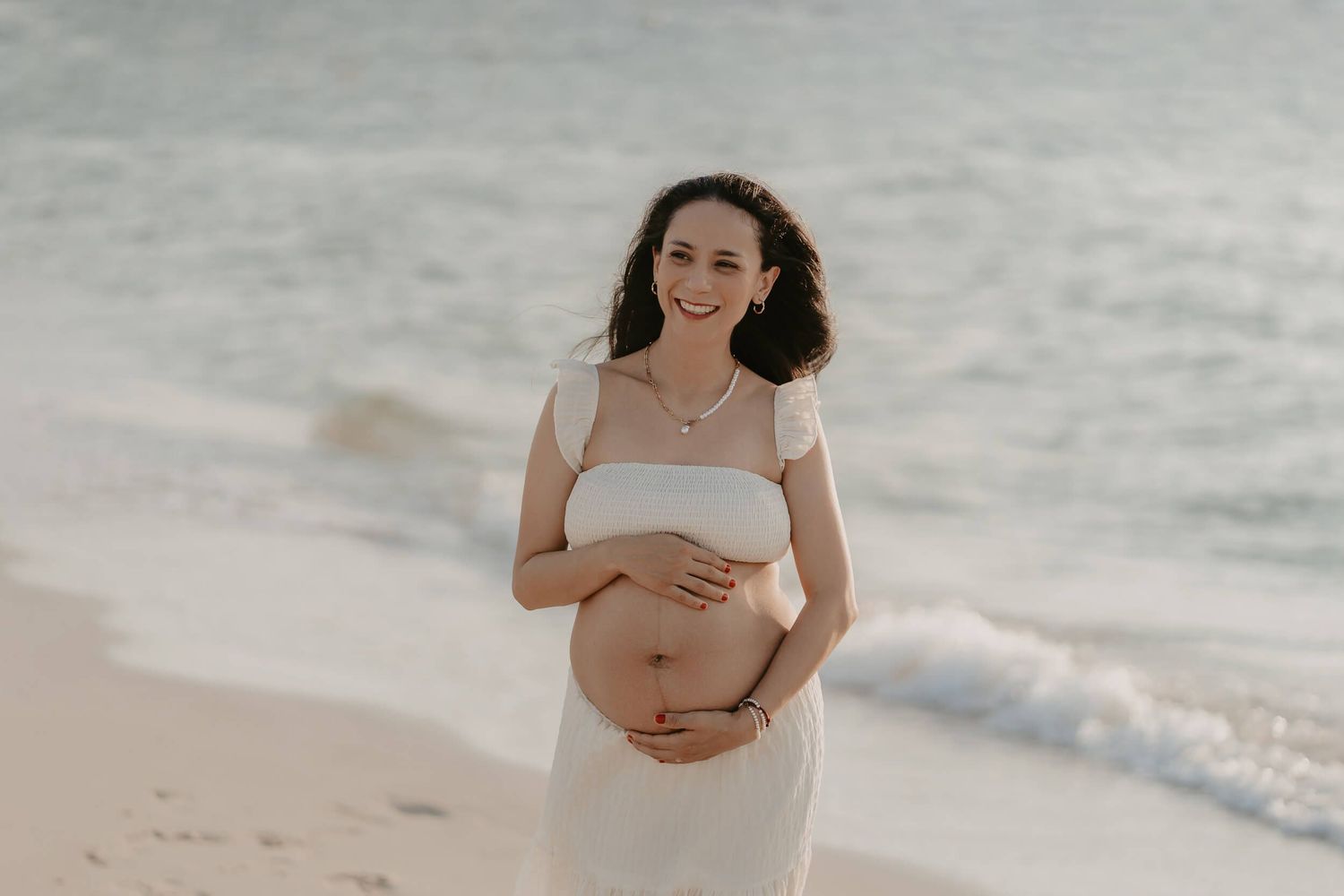 Expecting mother in a white dress holds her baby bump while standing on a beach at sunset.