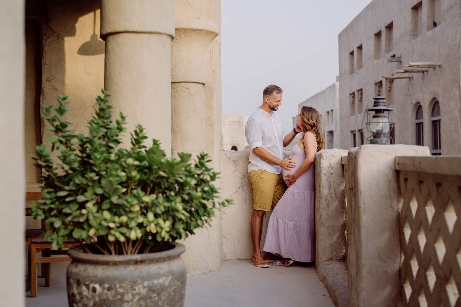 Two people share a romantic moment on a Mediterranean style balcony with stone columns and potted greenery.