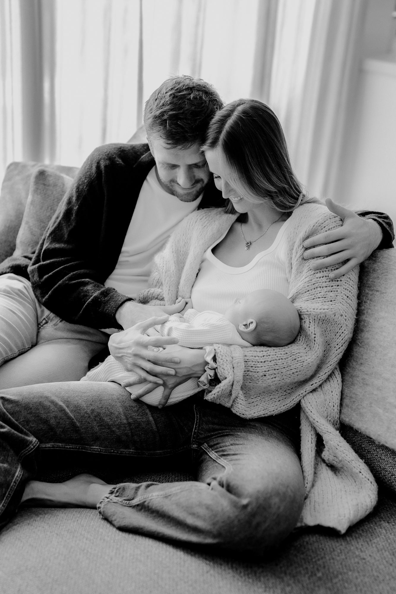 A tender black and white photograph captures a couple on a couch sharing an intimate moment with their newborn baby.