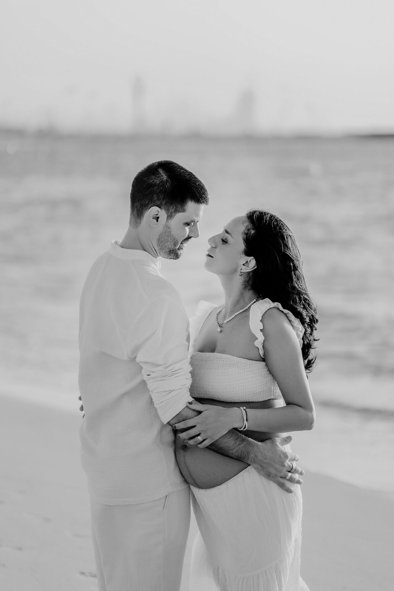 A tender black and white maternity photo of a couple embracing on a beach with ocean waves in the background.