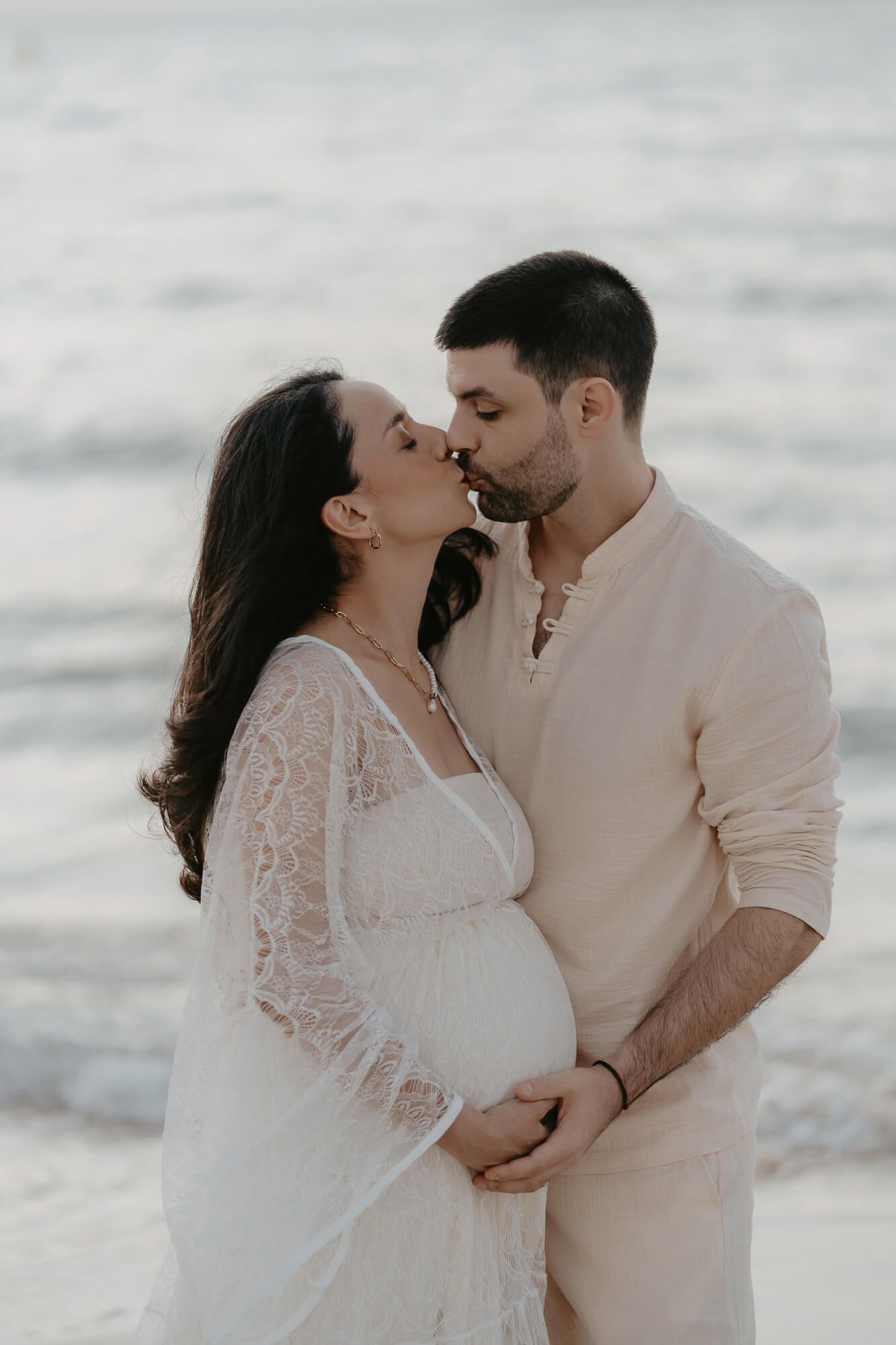 A romantic couple in light-colored attire shares an intimate moment by the ocean shore.