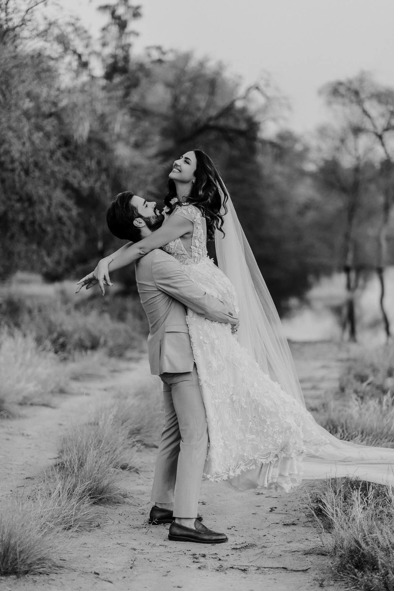 A newlywed couple dances together in a romantic desert setting with flowing veil and muted natural tones.
