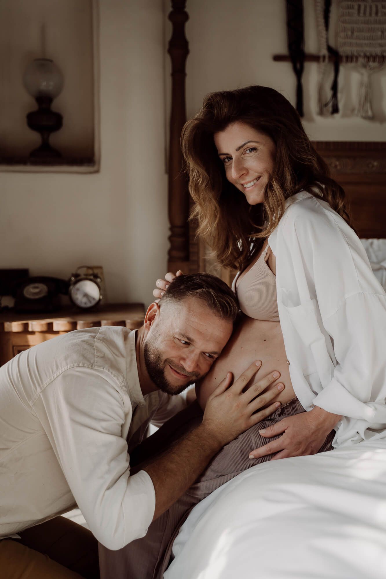 An intimate maternity photo showing expectant parents sharing a tender moment together on a white bed.