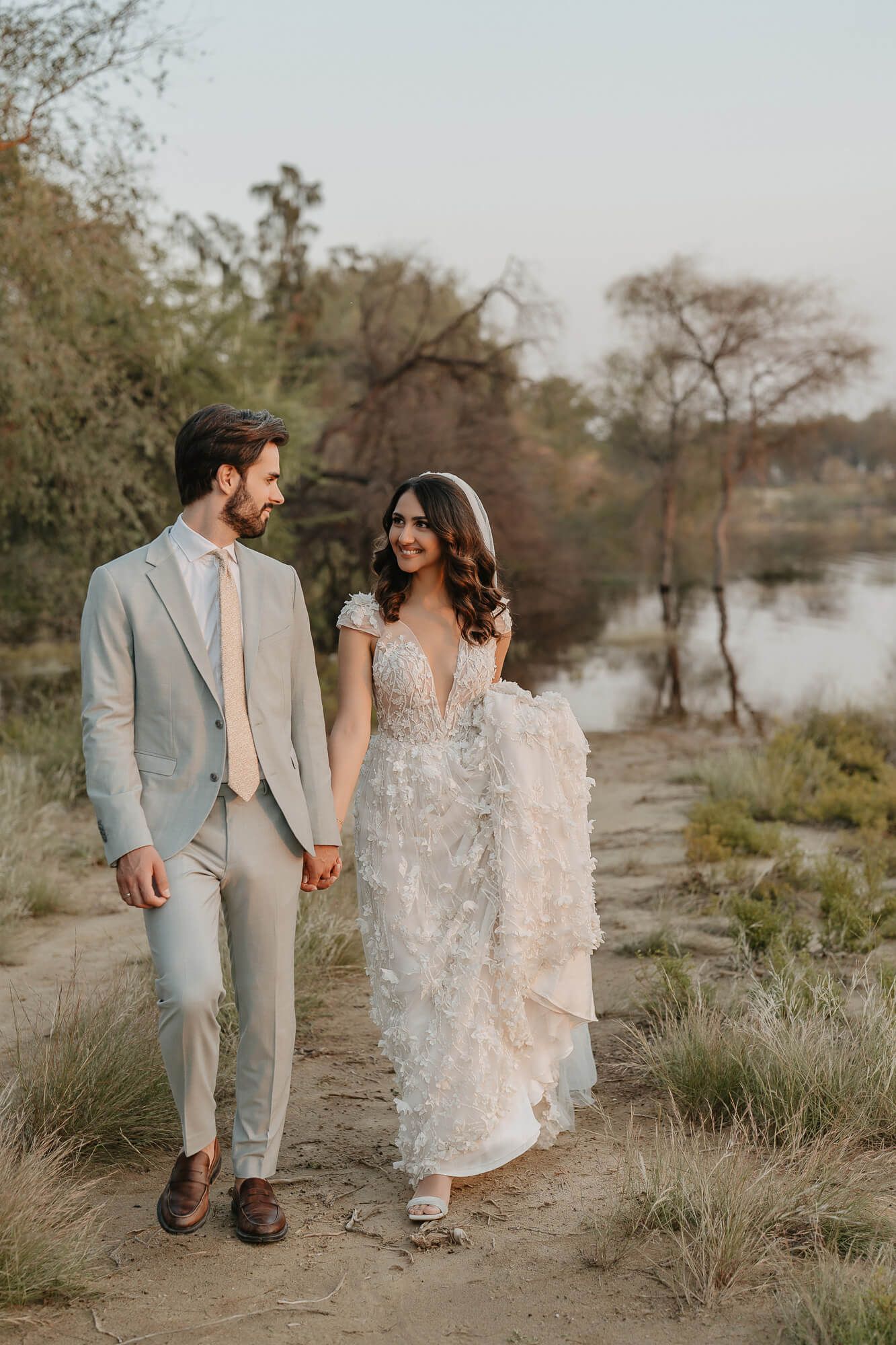 A romantic couple walking hand in hand by a lake, the bride wearing a flowing lace dress and groom in light gray suit.