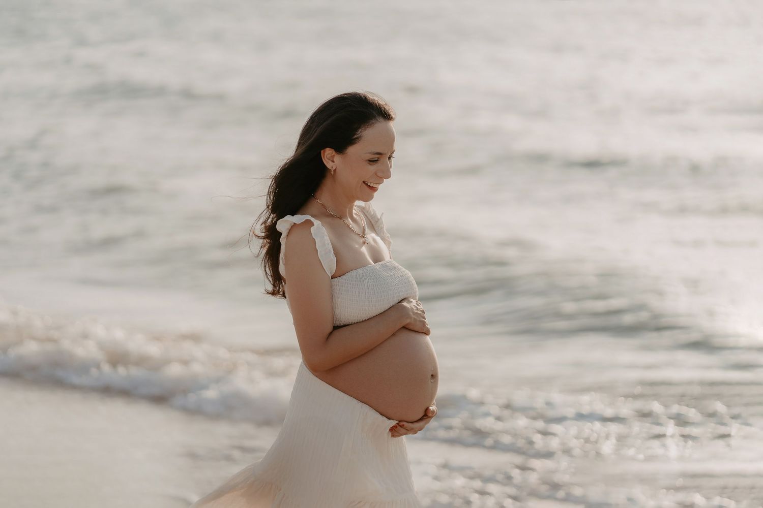 Expectant mother in a white dress stands peacefully on a beach with waves in the background.