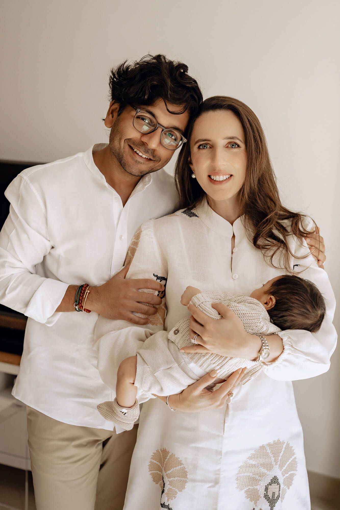 New parents in white clothing hold their infant baby while posing together for a heartwarming family portrait.