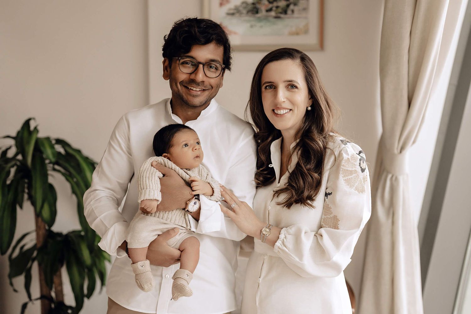 Young couple in white outfits hold their infant baby in a cozy indoor setting with plants and artwork.