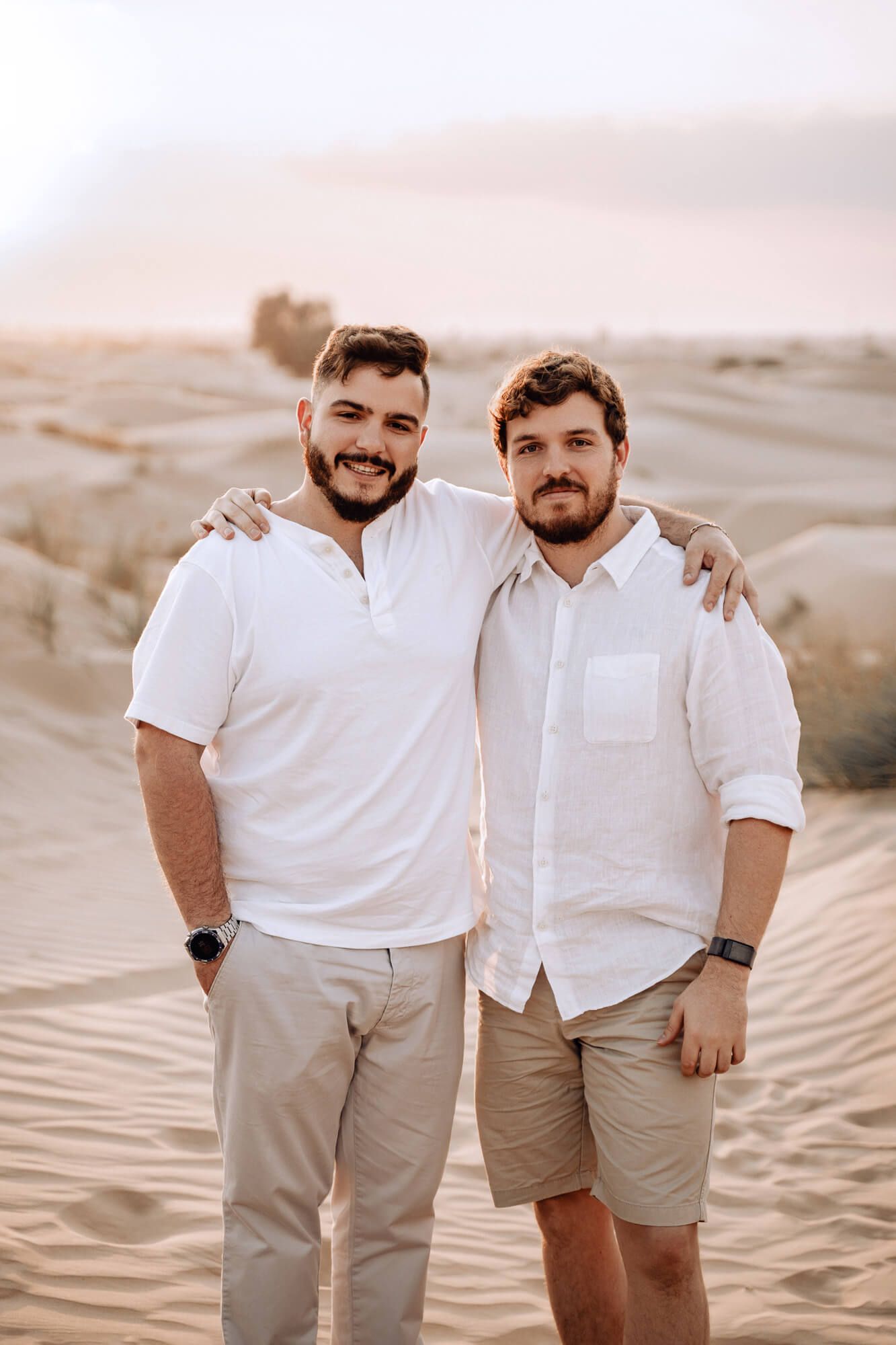 Two people in coordinated white shirts and khaki clothing pose together on desert sand dunes.