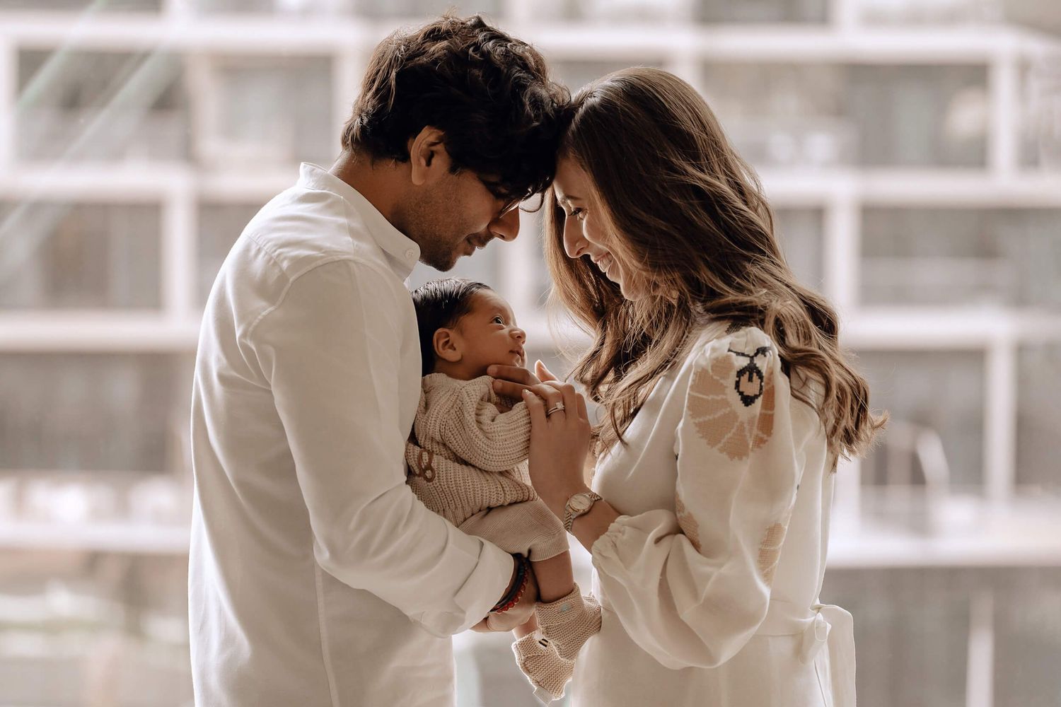 Intimate family portrait with parents in white embracing their baby in soft window light.