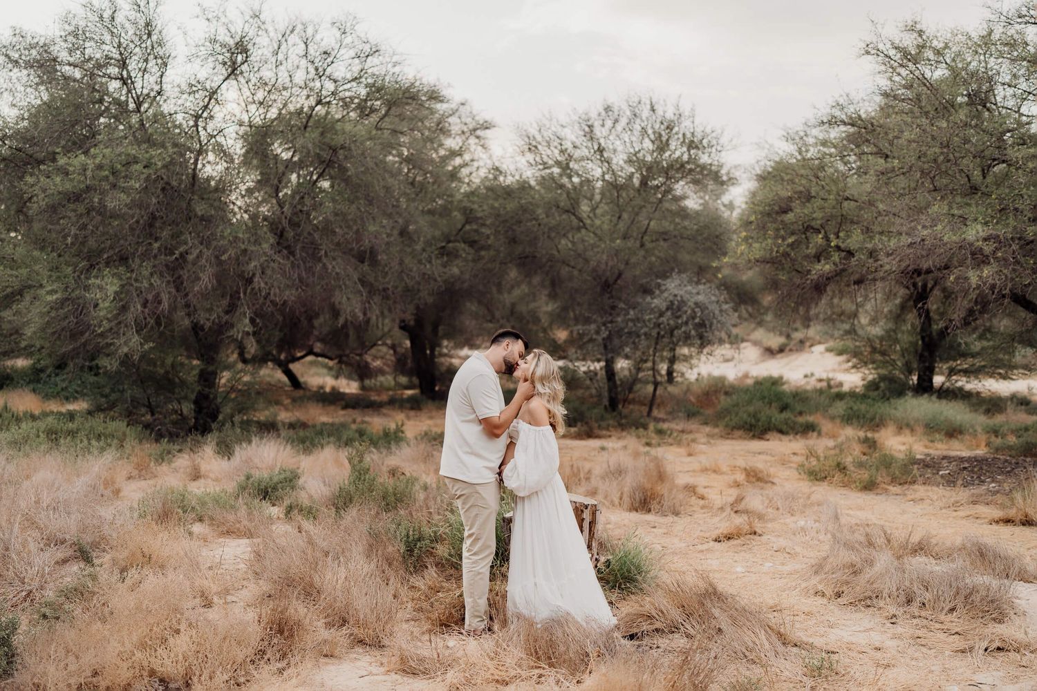 Desert maternity session with couple in neutral tones surrounded by sparse trees.