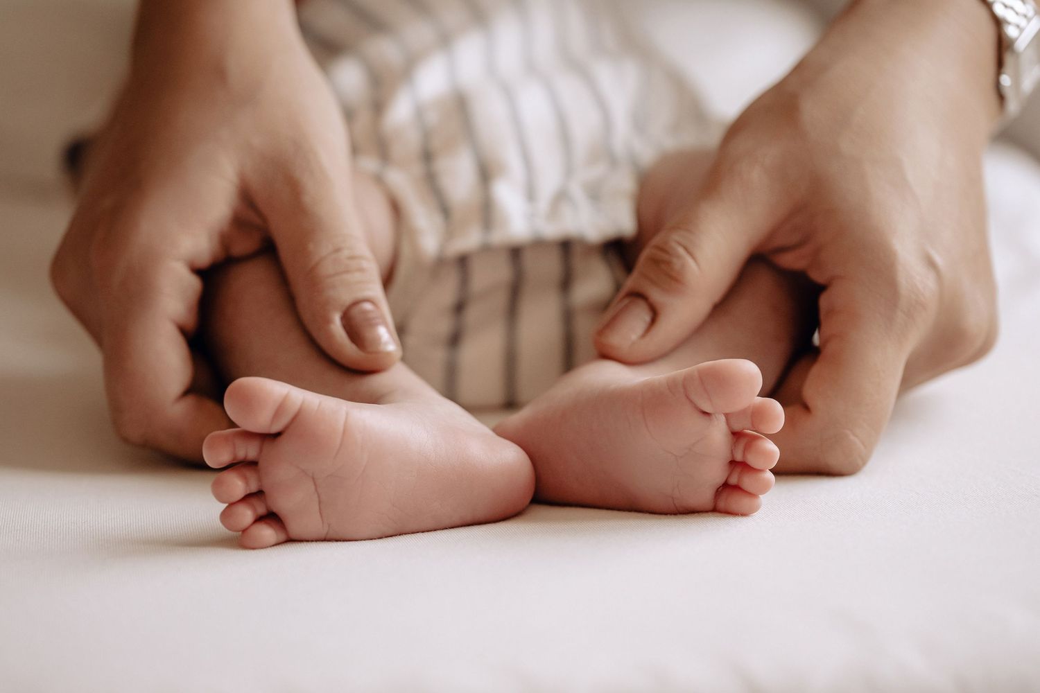 Adult hands gently touching tiny newborn baby feet on white bedding.