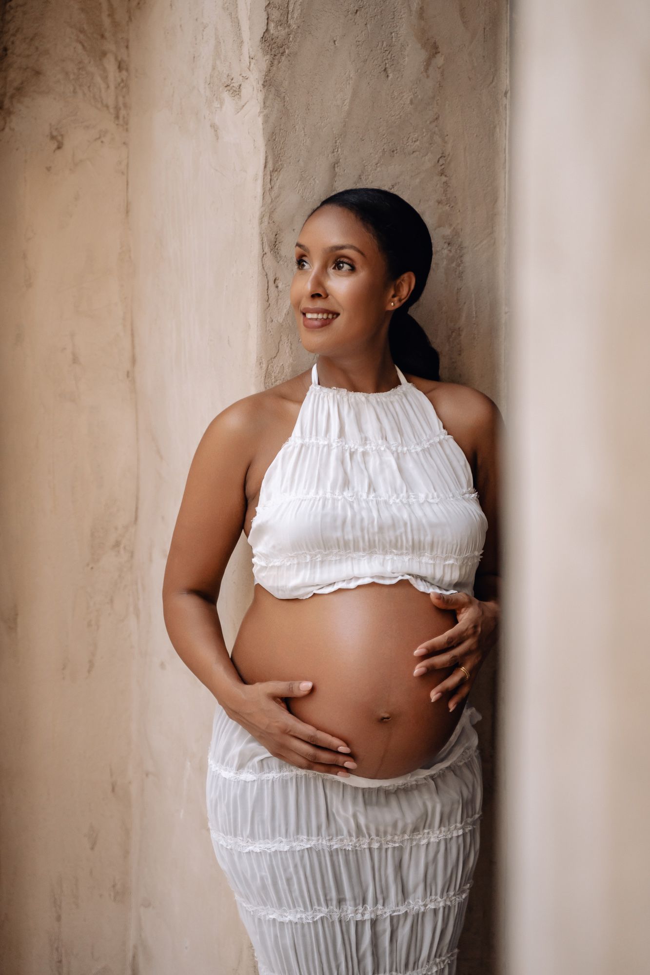 Expectant mother in a white ruched dress poses against a textured wall during a maternity photoshoot.