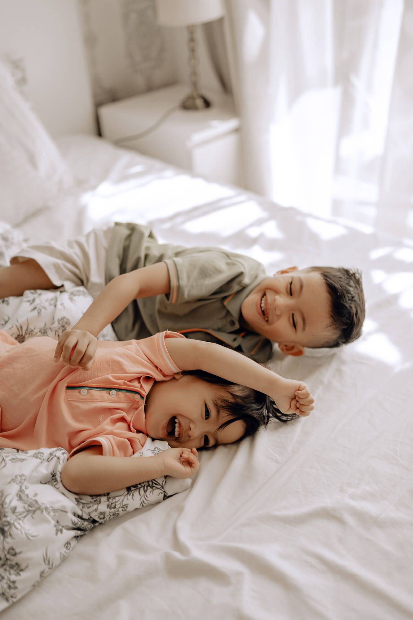 Two children laughing while lying on white bedding in a bright, sunlit bedroom.