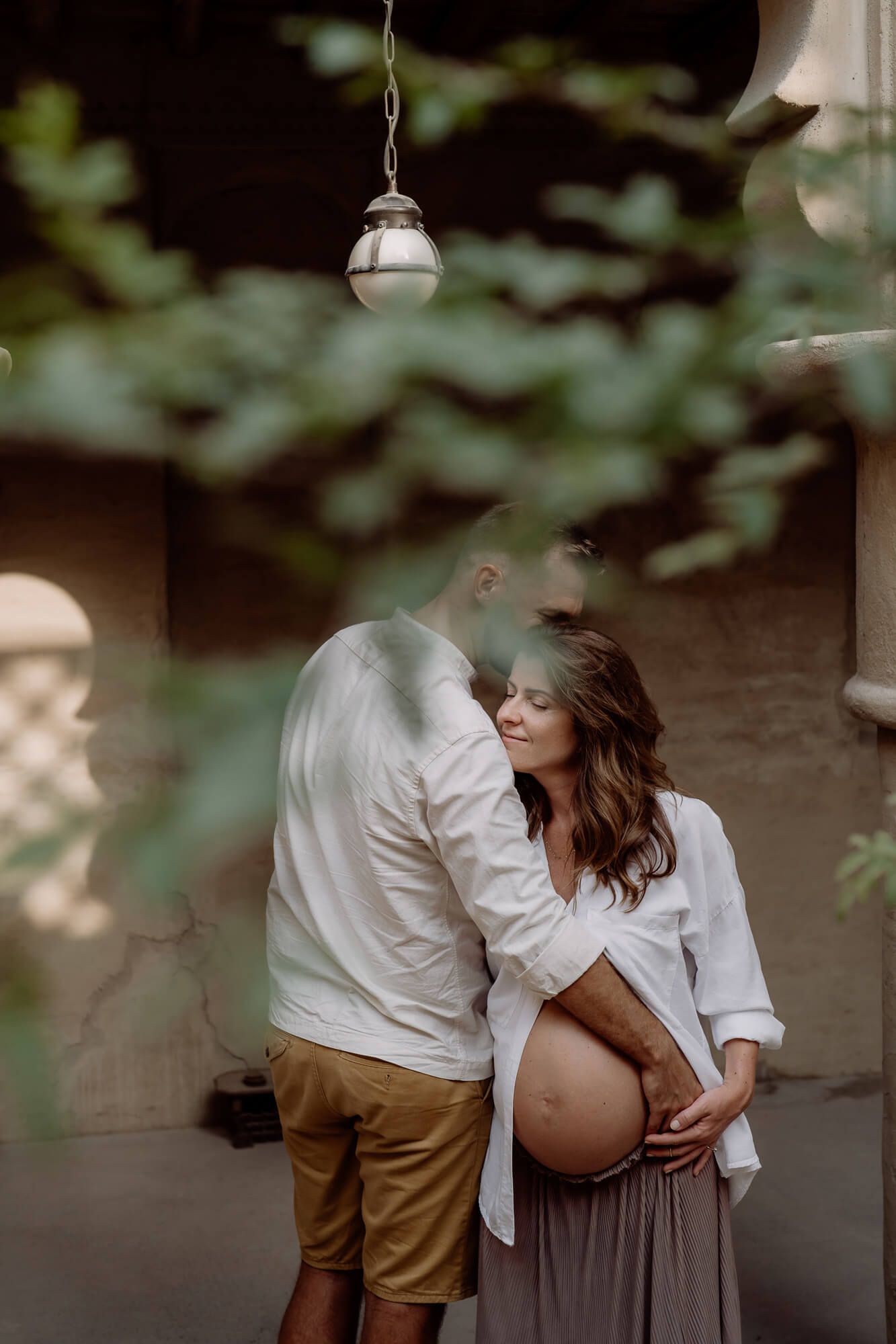 A couple shares an intimate moment together outdoors wearing white and beige clothing in dappled sunlight.